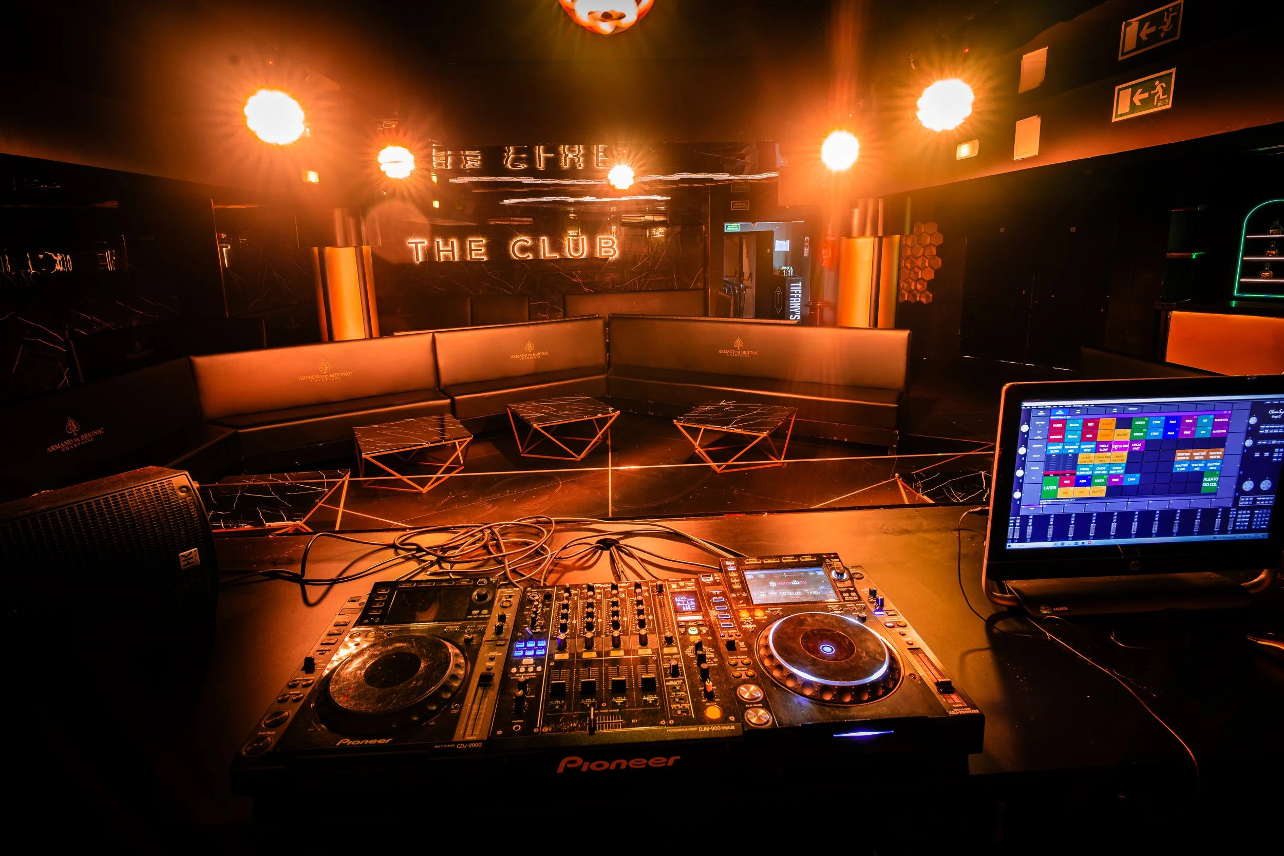 Interior de una discoteca con luces anaranjadas y un cartel que dice "THE CLUB" en la pared del fondo. En el frente hay una mesa con equipo de DJ, incluyendo tornamesas y consola de mezclas. A la derecha, una laptop con una pantalla que muestra un pr