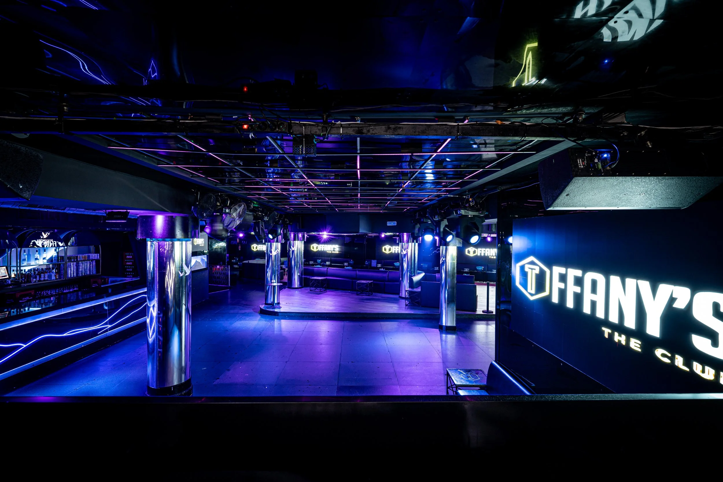 Interior de un club nocturno con iluminación en tonos violetas y azules, barras y equipo de DJ, señalización luminosa que dice 'T FANY'S THE CLUB'.