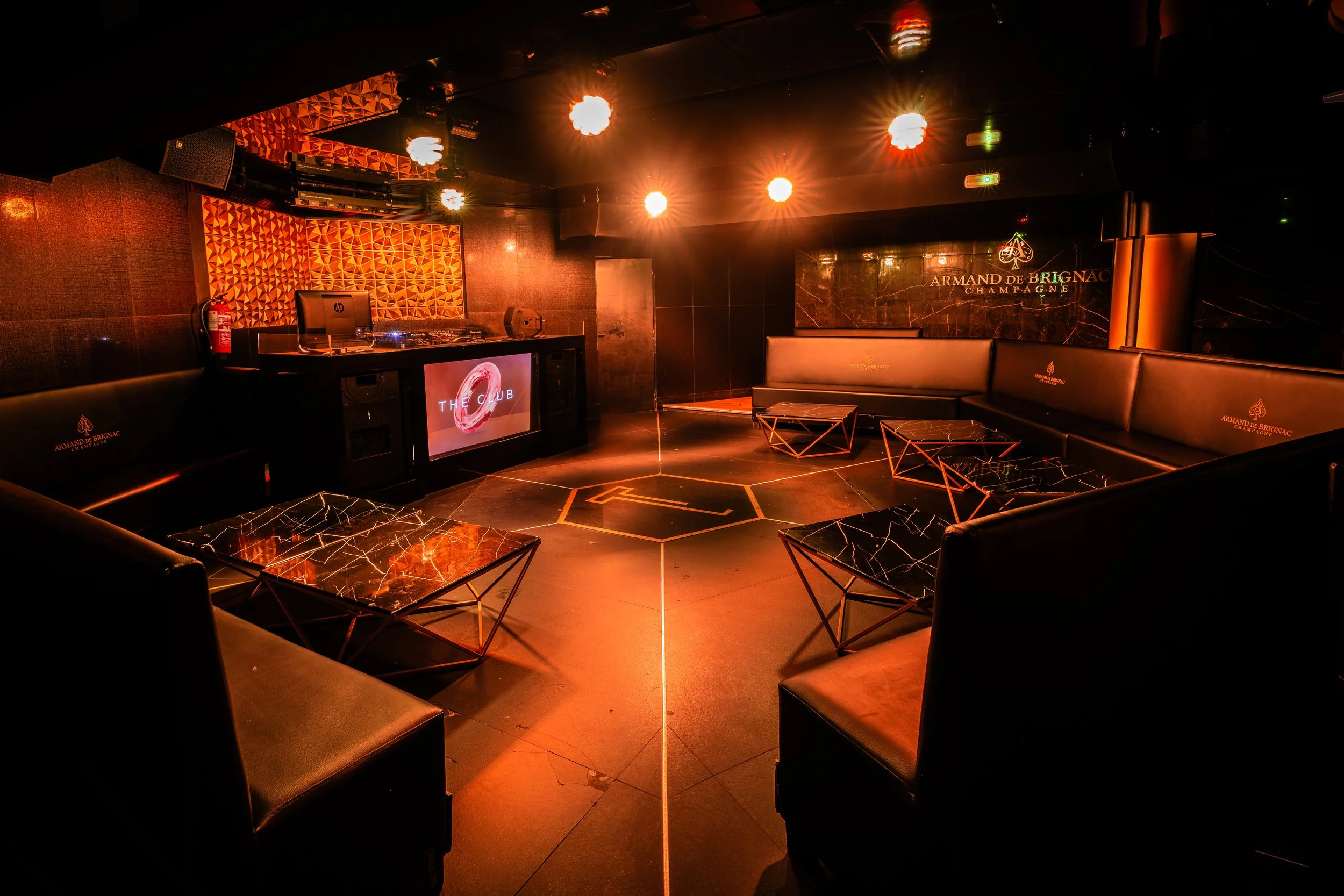 Interior de un club nocturno con luces anaranjadas, área de asientos con sillones negros y mesas de mármol, una pared con logotipos de Armand de Brignac Champagne y una pared con una pantalla de DJ. Ambiente elegante y moderno.