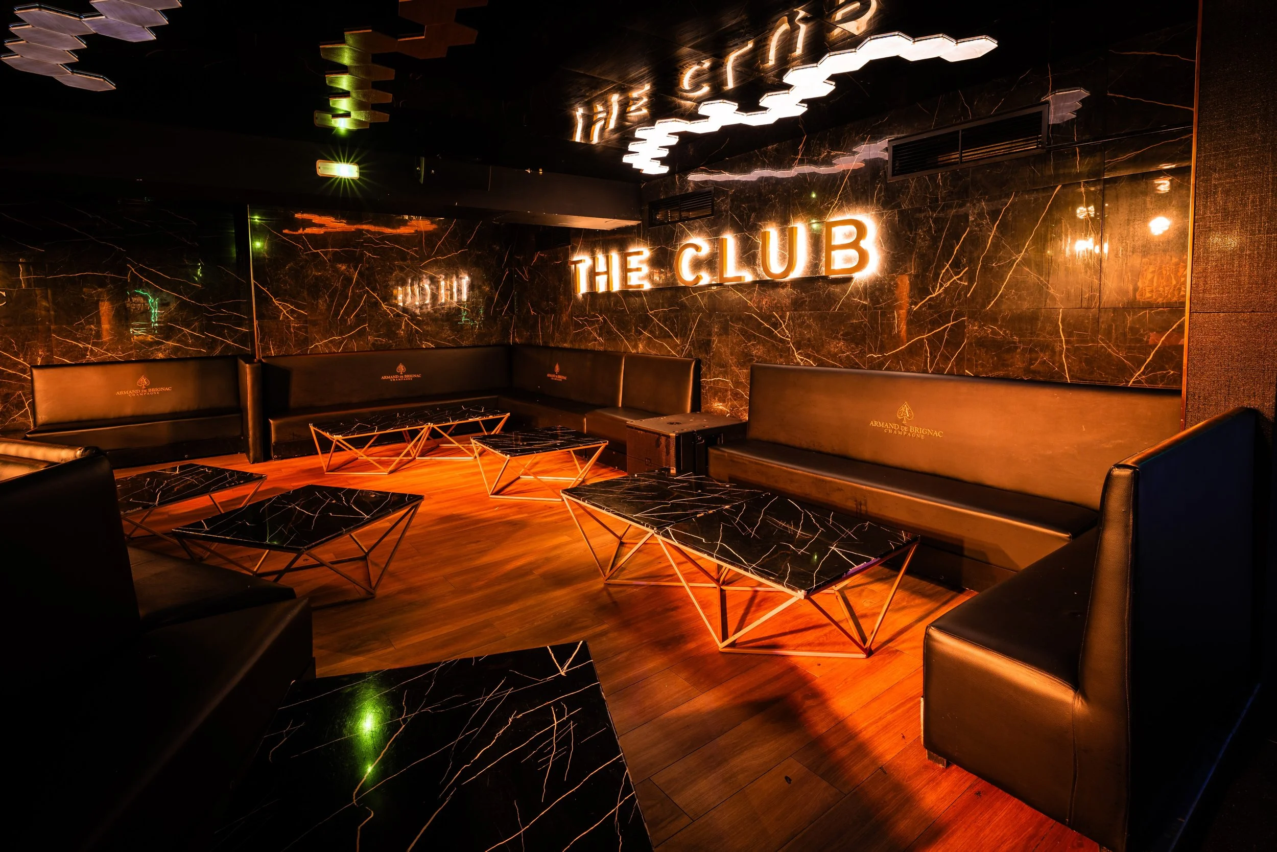 Interior de un club nocturno con paredes de mármol negro y neón naranja que dice 'THE CLUB'. Muebles de asiento de cuero negro y mesas de mármol negro con patas doradas, piso de madera y decoración moderna con luces LED.