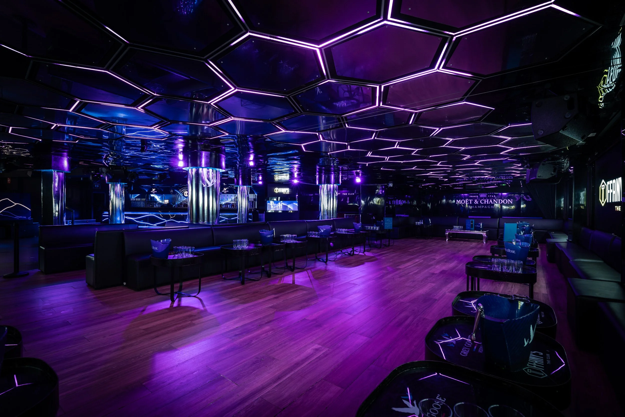 Interior de un club nocturno con iluminación en tonos morados y violetas, techo de hexágonos con luces LED, sillones y mesas, ambiente moderno y cosmopolita.