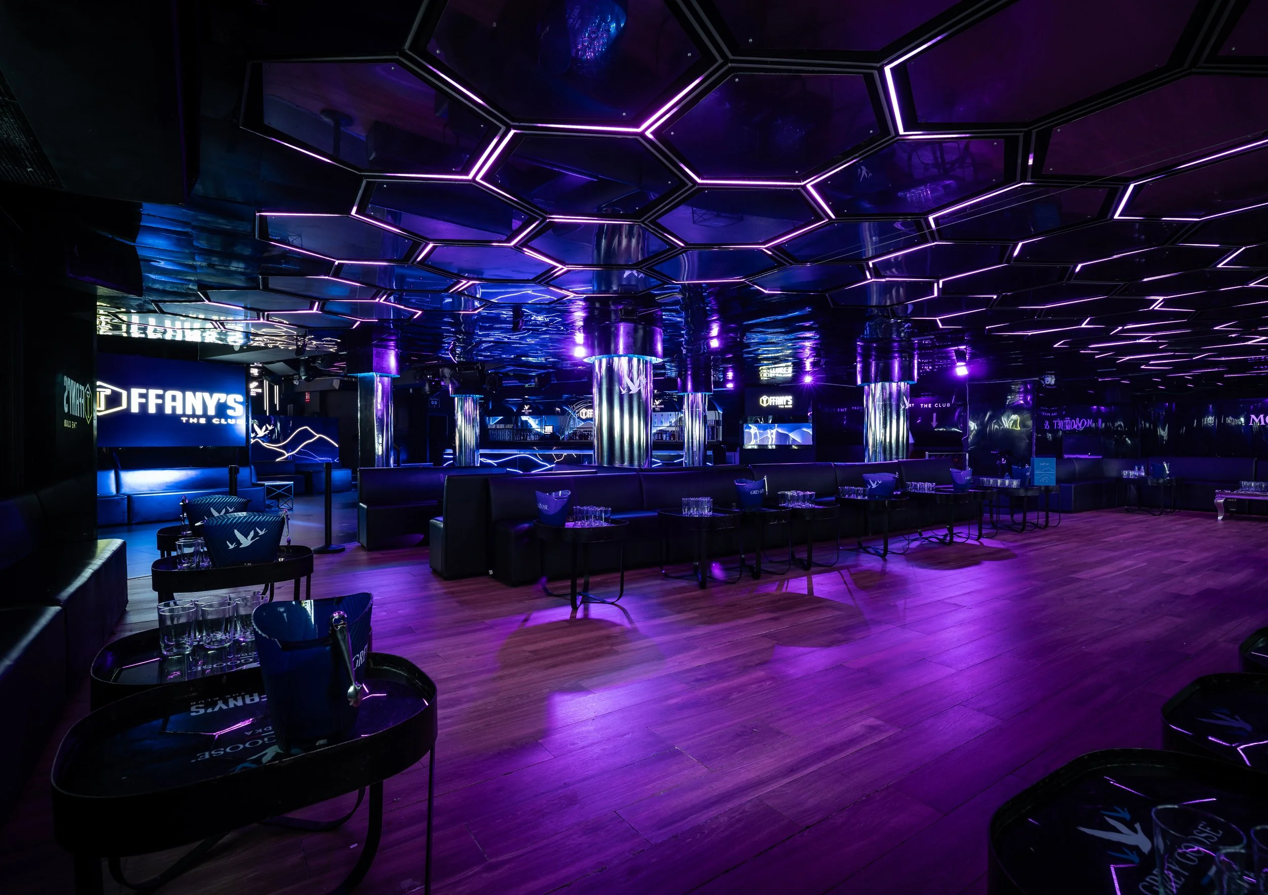 Interior de un club nocturno con iluminación azul y púrpura, mesas con vasos y sillas, techo con luces de neón en patrones hexagonales, columnas de metal y pantallas electrónicas.