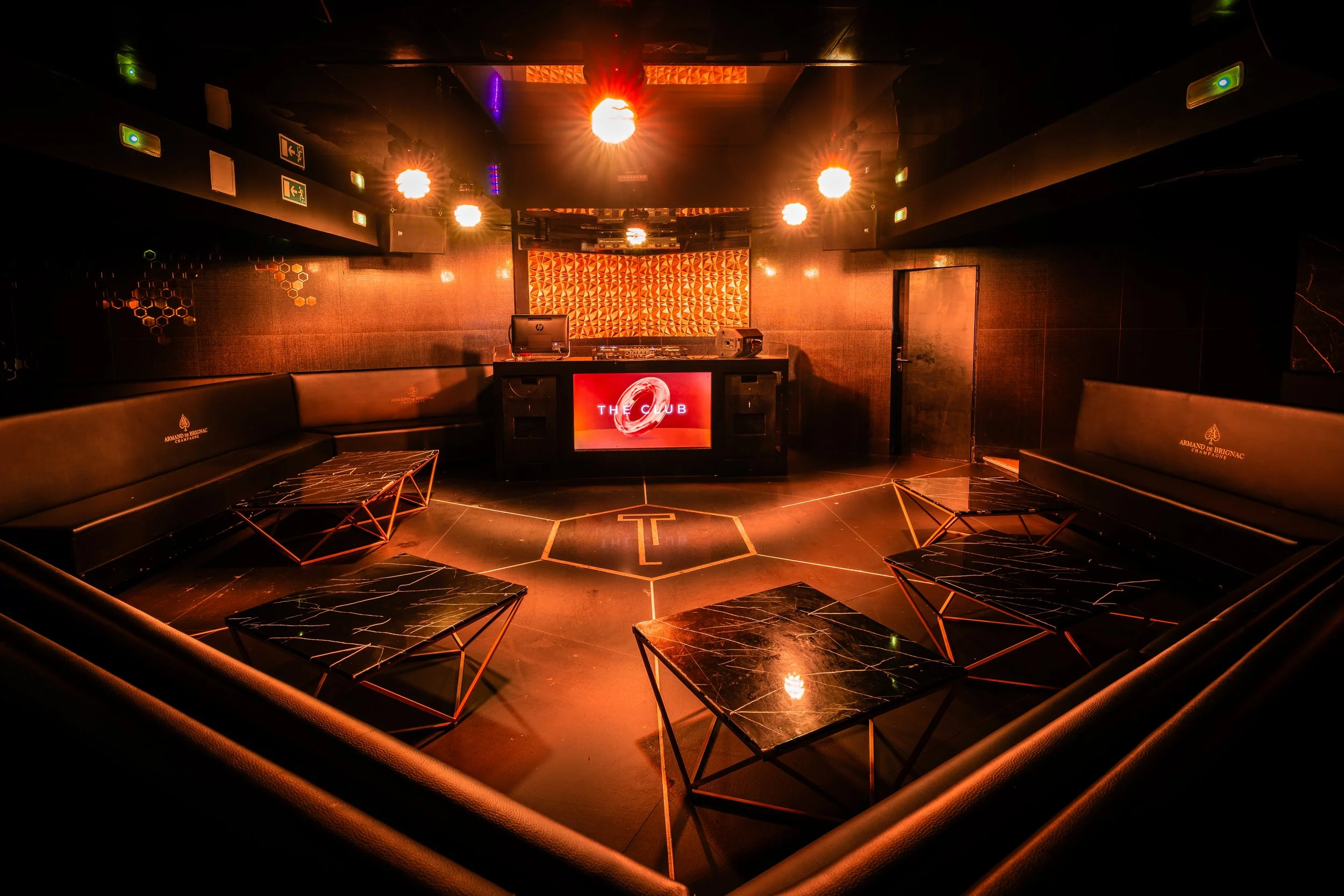 Interior de un club nocturno con luces cálidas y mobiliario moderno, incluyendo bancos de cuero y mesas de mármol negro con patrones blancos, en el escenario hay un equipo de DJ y una pantalla que dice 'The Club'.