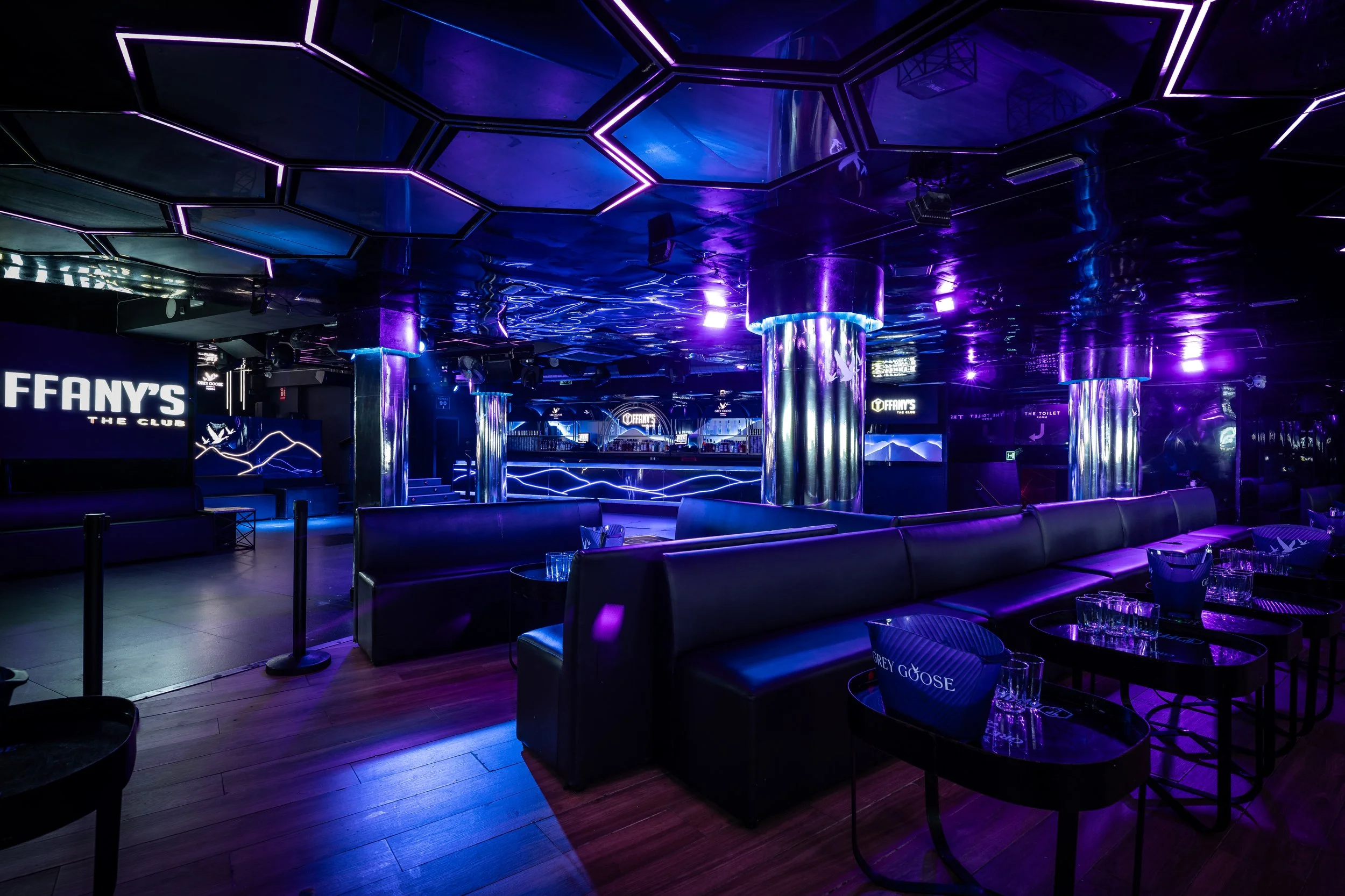 Interior de club nocturno con iluminación neon púrpura y cromo, mesas y sillas de cuero negro, pantallas y barras en el fondo.