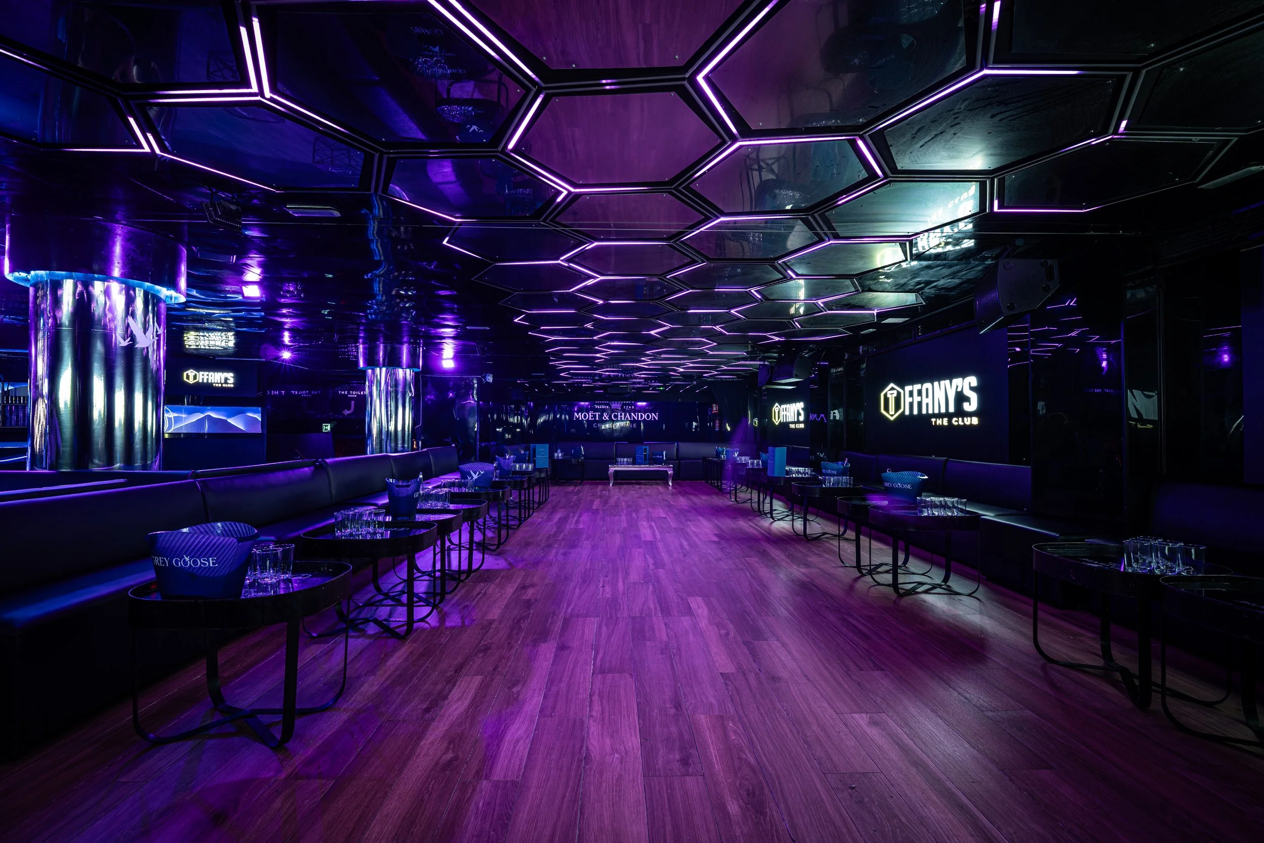 Interior de un club nocturno con iluminación púrpura, mesas con vasos y sillas, paredes decoradas con pantallas y señalización que dice 'FANY'S THE CLUB'.