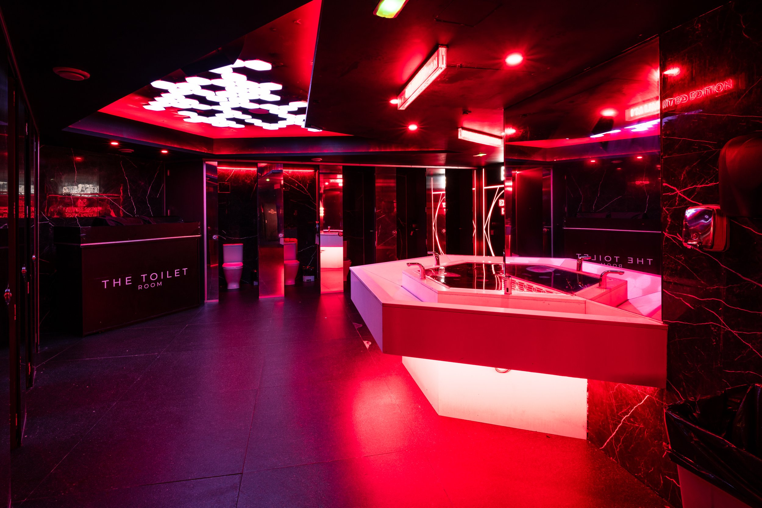 Interior de un baño público con iluminación en tonos rojos y detalles en mármol negro, hay un área con lavabo y un cartel que dice 'The Toilet Room'.