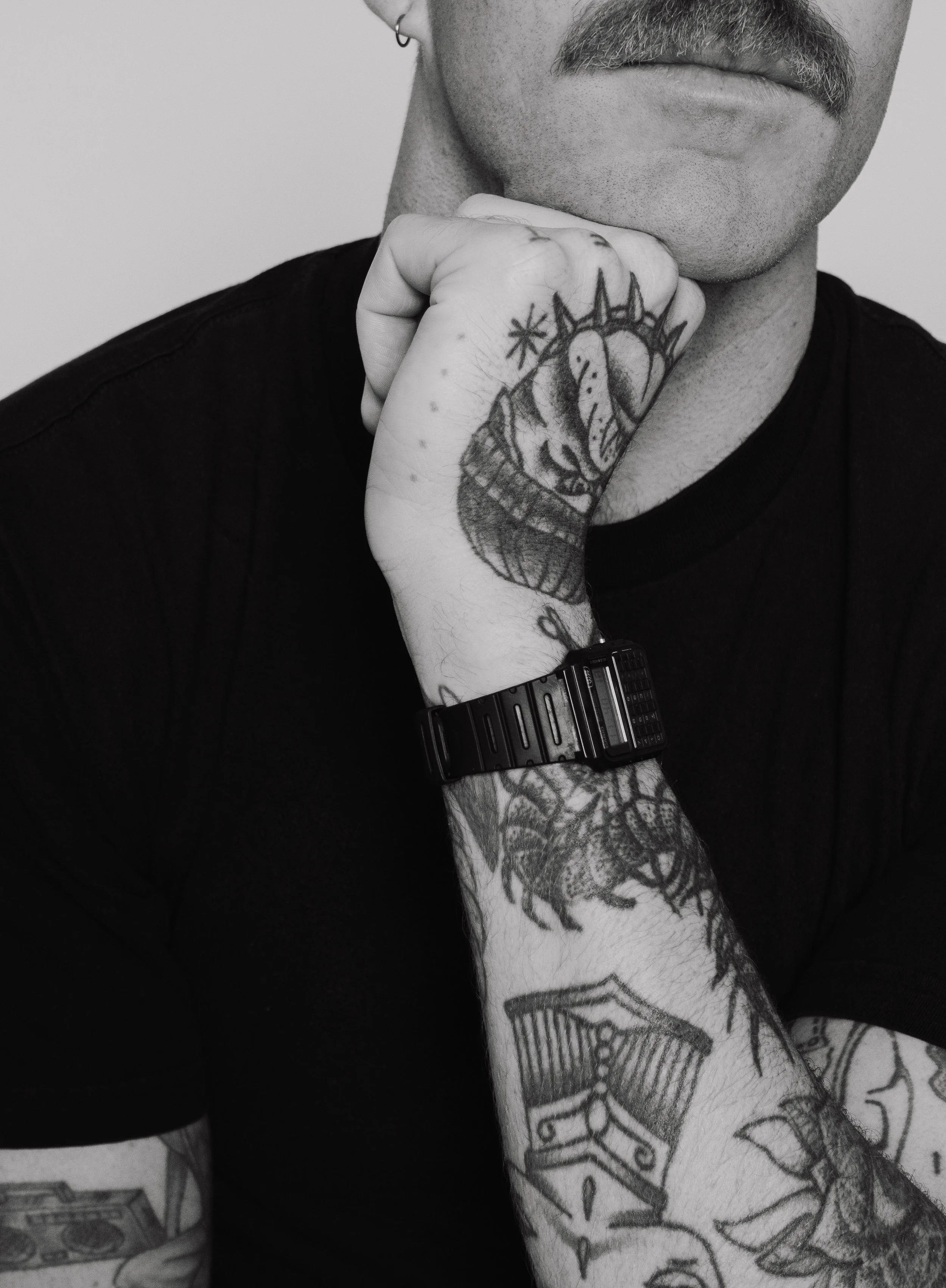 Close-up of a man's face and arm with tattoos, wearing a black T-shirt and black watch, resting his chin on his hand.