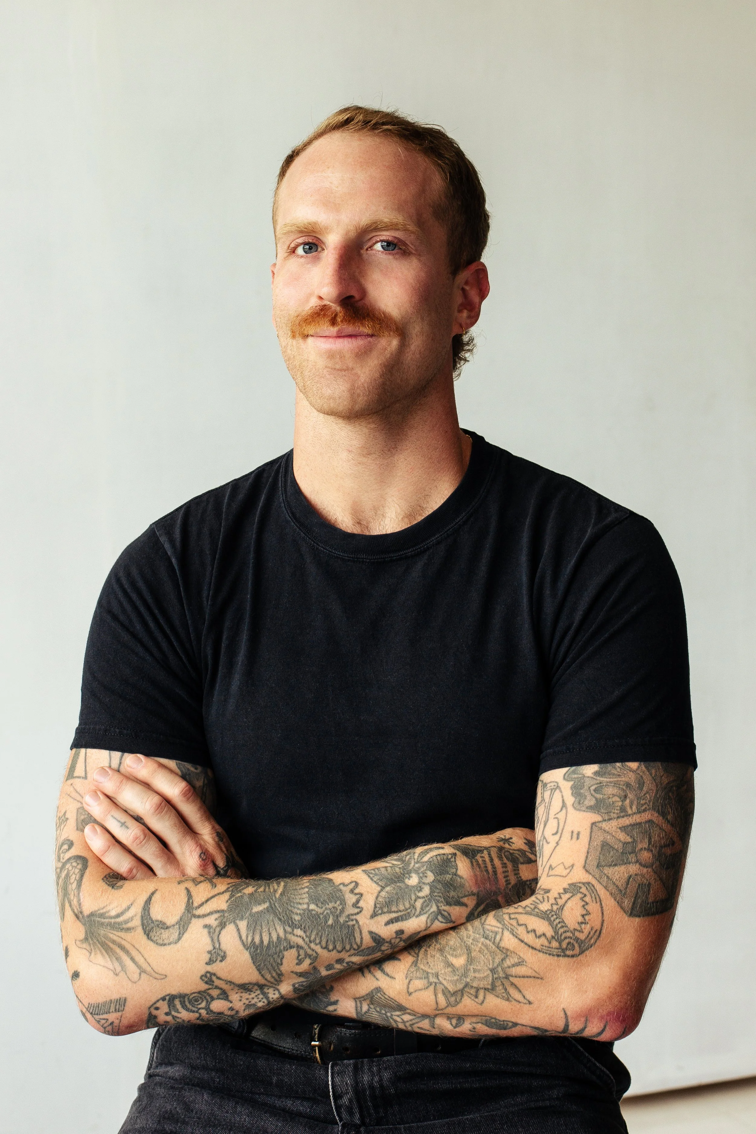 A man with tattoos on his arms and a mustache, wearing a black t-shirt, standing with his arms crossed against a plain light-colored background.