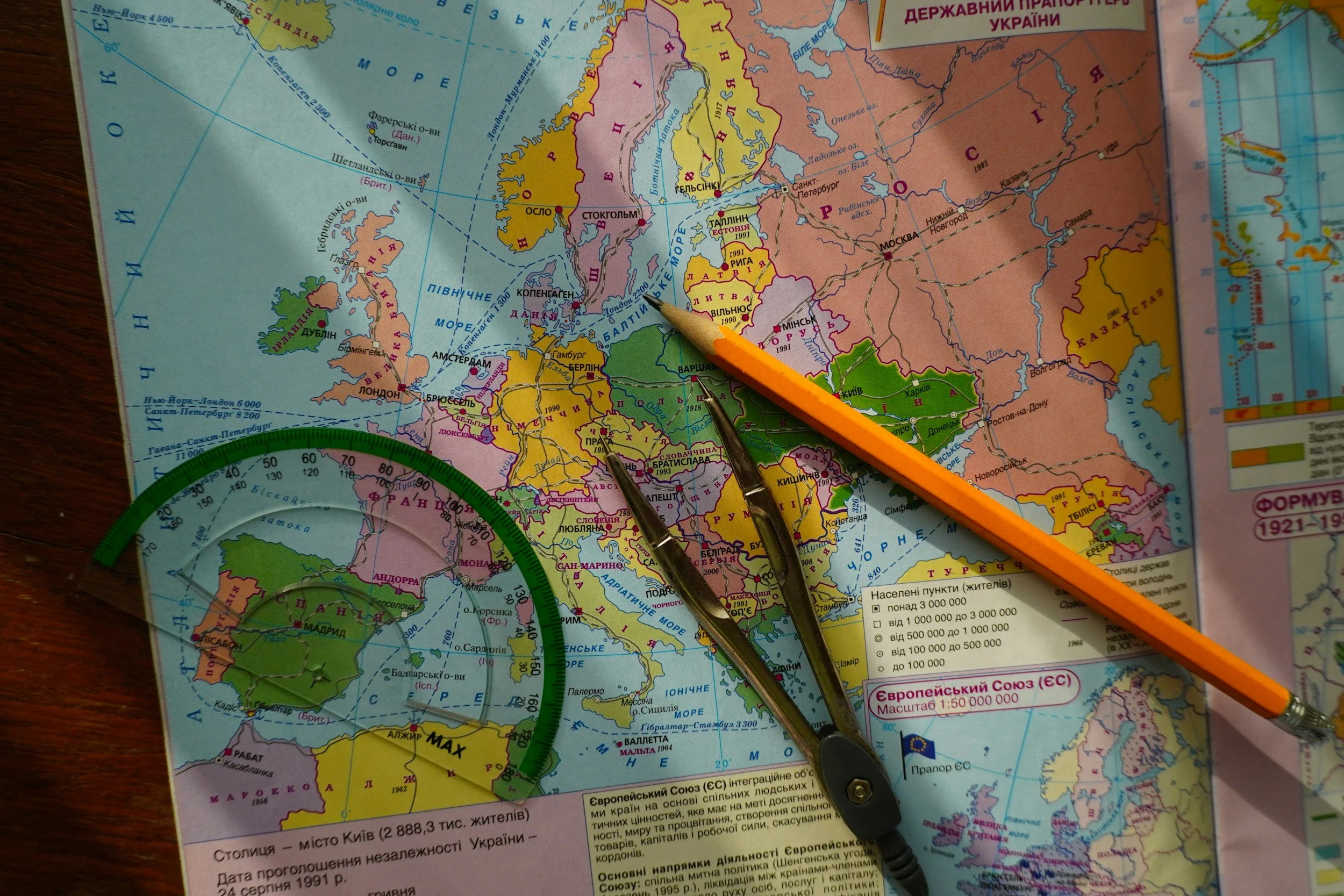 A map of Europe with various countries color-coded. There are several drawing tools like a pencil, a pair of compasses, and a protractor placed on the map. The map includes country names, some city names, and other geographical details in Ukrainian.