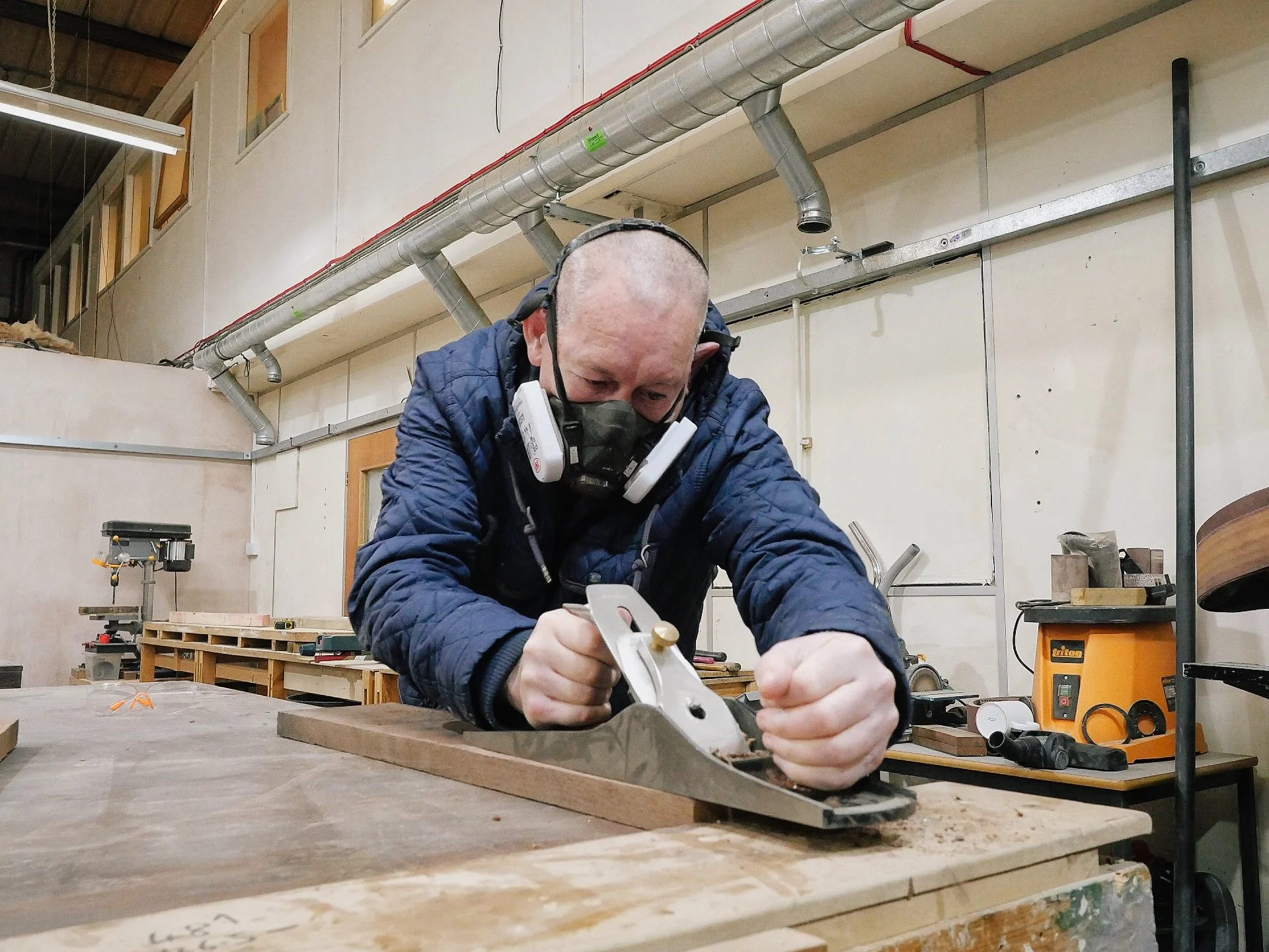 A male service user with a learning disability wearing a protective mask and glasses is working on a woodworking project in the Key workshop. He is using a handheld power tool on a piece of wood.