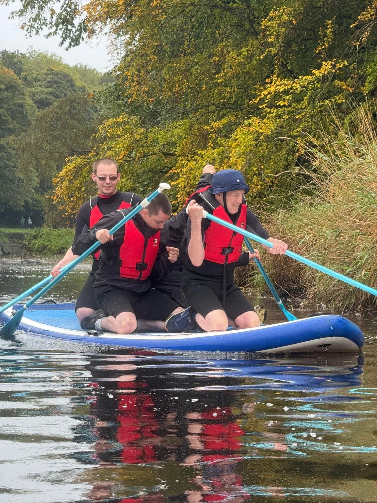 Three service users with learning disabilities in life jackets and wetsuits, kneeling on a paddleboard in a river, holding paddles, with trees and foliage in the background.