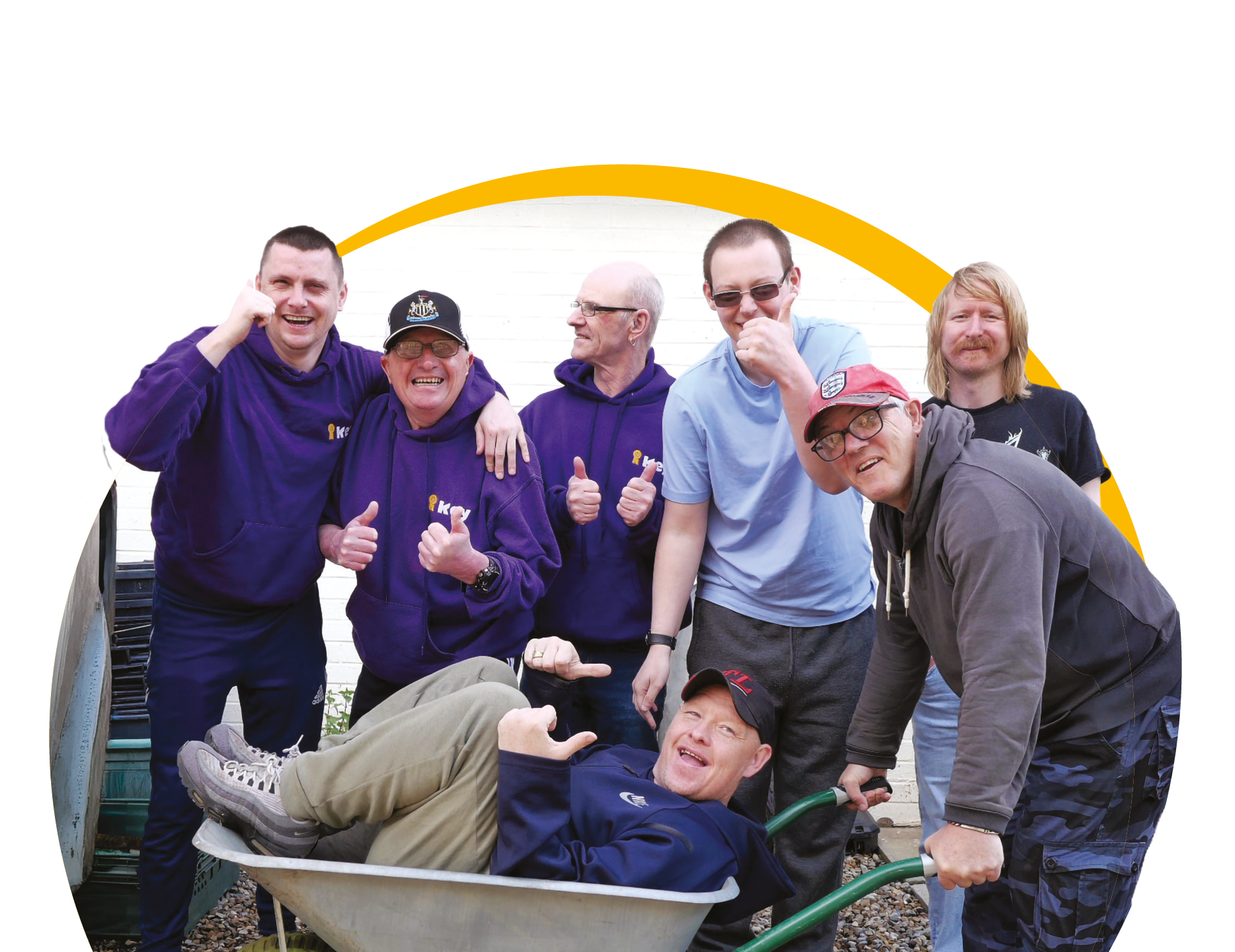 Group of seven service users with learning disabilities posing happily for a photo, some giving thumbs up, one lying in a wheelbarrow, outdoors in front of the Key garden.