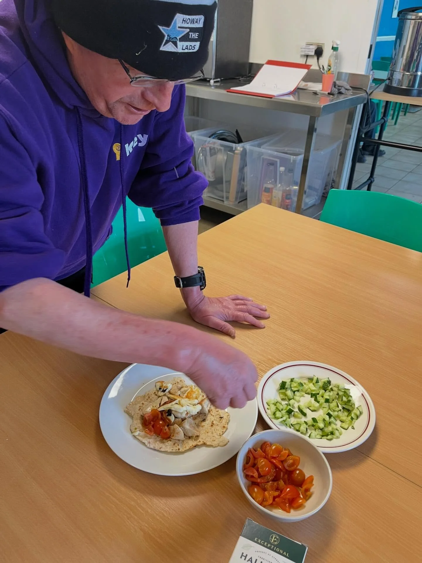 A service user wearing a purple Key merch hoodie and a black Newcastle hat prepares a meal at a dining table in the canteen at Key.
