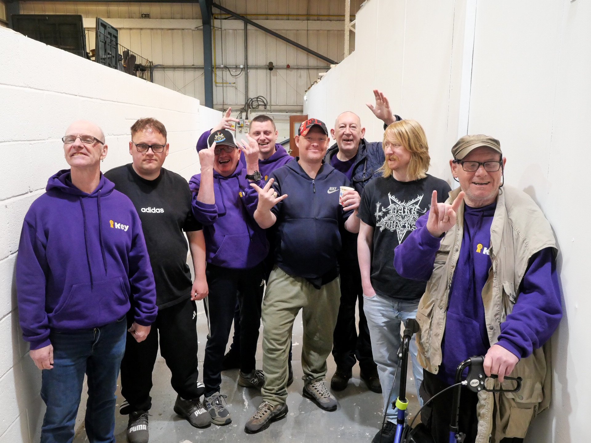 A group of service users, all men with learning disabilities stand, posing for a photo, some making sillyhand gestures, one person with mobility aid, dressed casually Key merch hoodies and t-shirts.