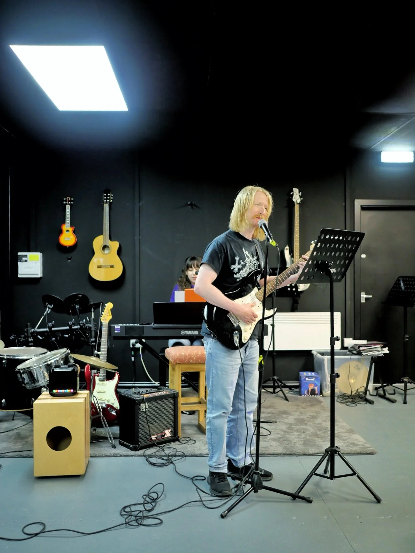 A service user with autism playing an electric guitar and singing into a microphone in Key's black box studio space. Behind him are guitars mounted on the wall, and a music therapist sits behind accompanying him on the keyboard.