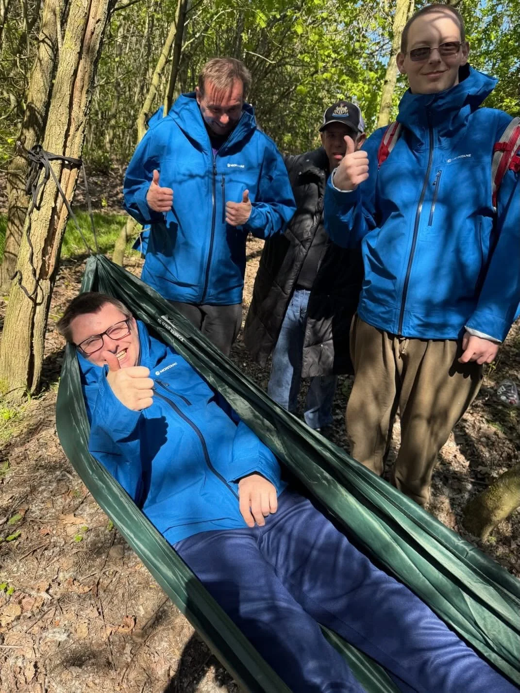 Four male service users in a wooded area, with one lying in a hammock giving a thumbs up and the others standing around smiling and giving thumbs up, wearing outdoor jackets.