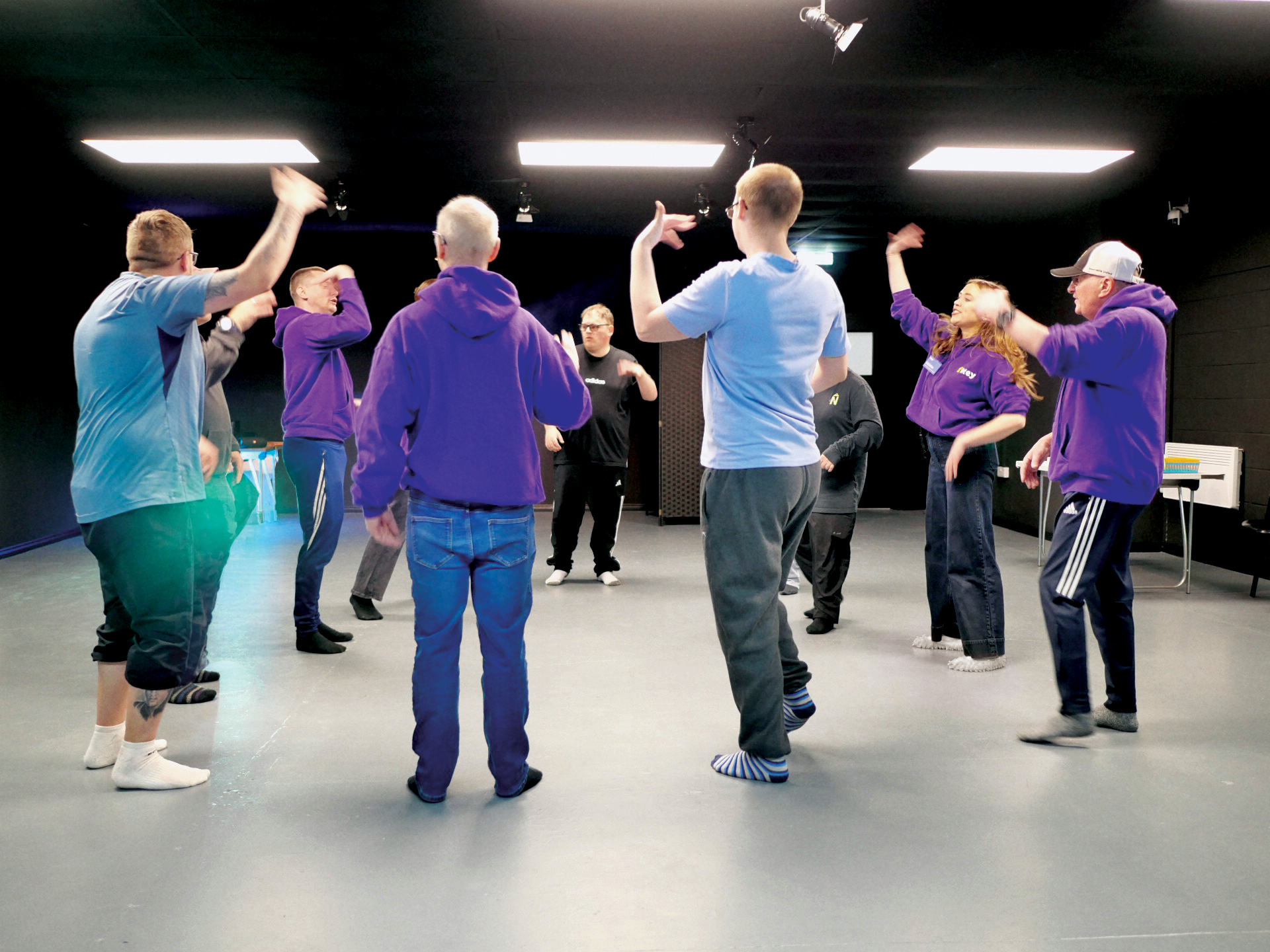 A group of service users with learning disabilities and acquired brain injuries participating in dance and drama in a black box studio at Key, most wearing casual and comfortable clothing with some in purple Key merch hoodies.