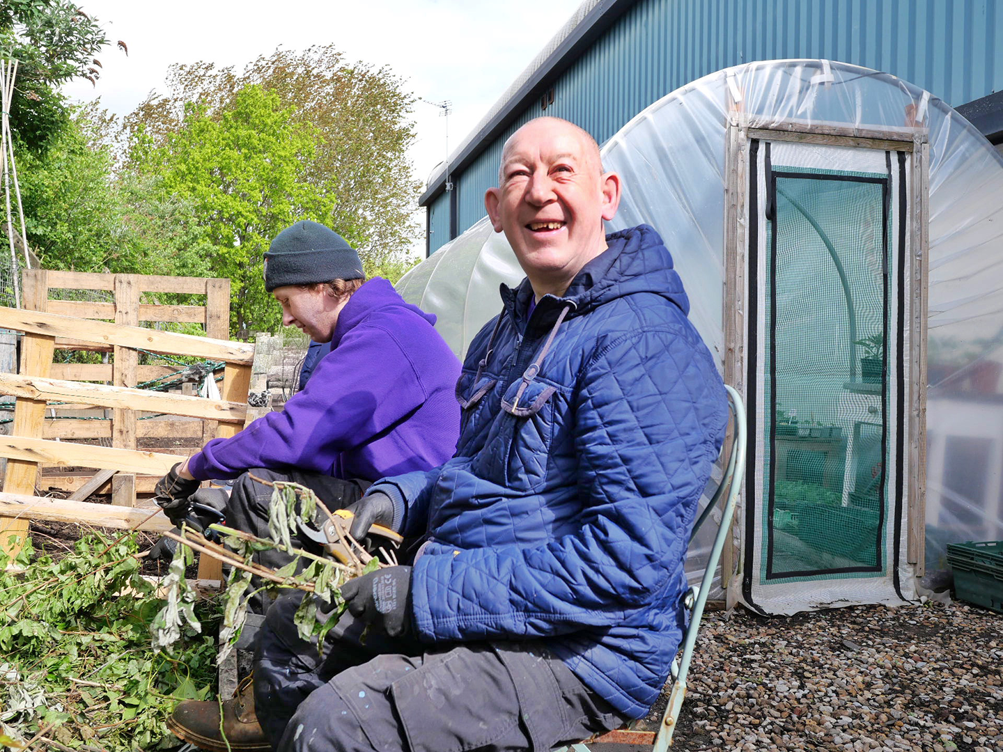 A service user, a man with a learning disability, sits in the garden doing some planting. He smiles at the camera and is sat next to a member of Key staff who is supporting him.