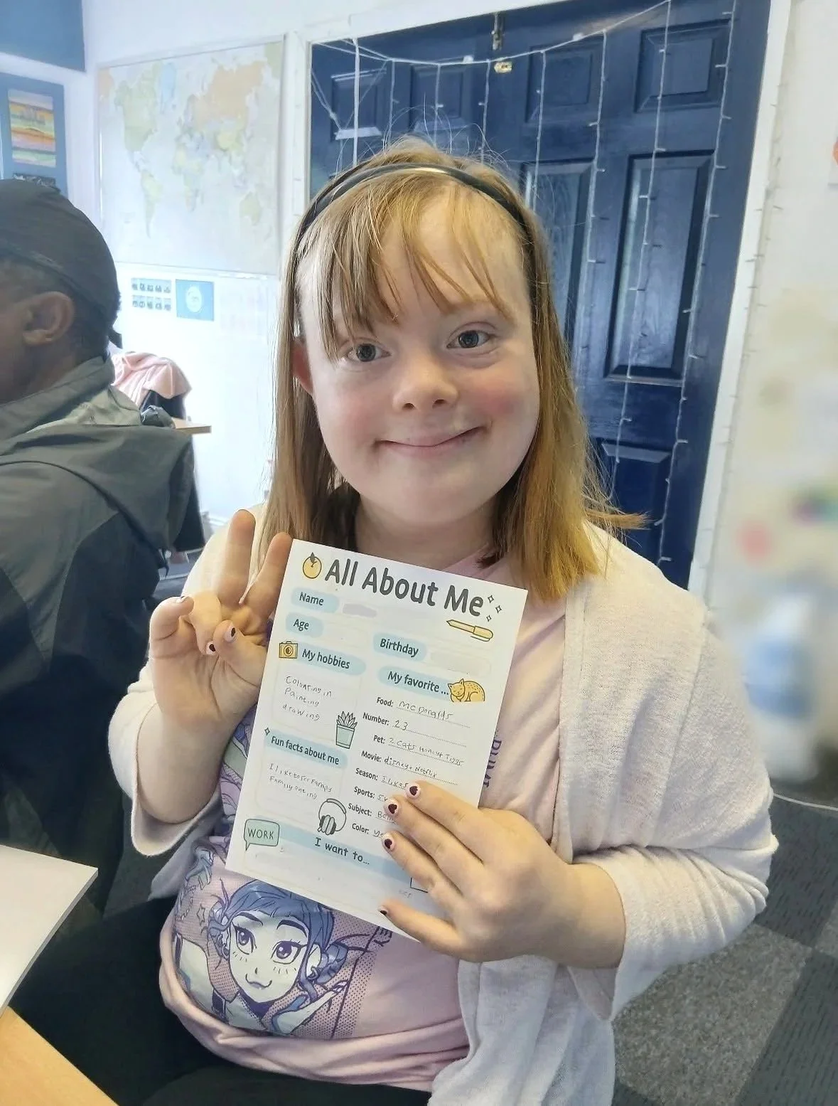A female service user with a learning disability smiling and holding an "All About Me" worksheet the Key classroom. She is making a peace sign with her right hand.