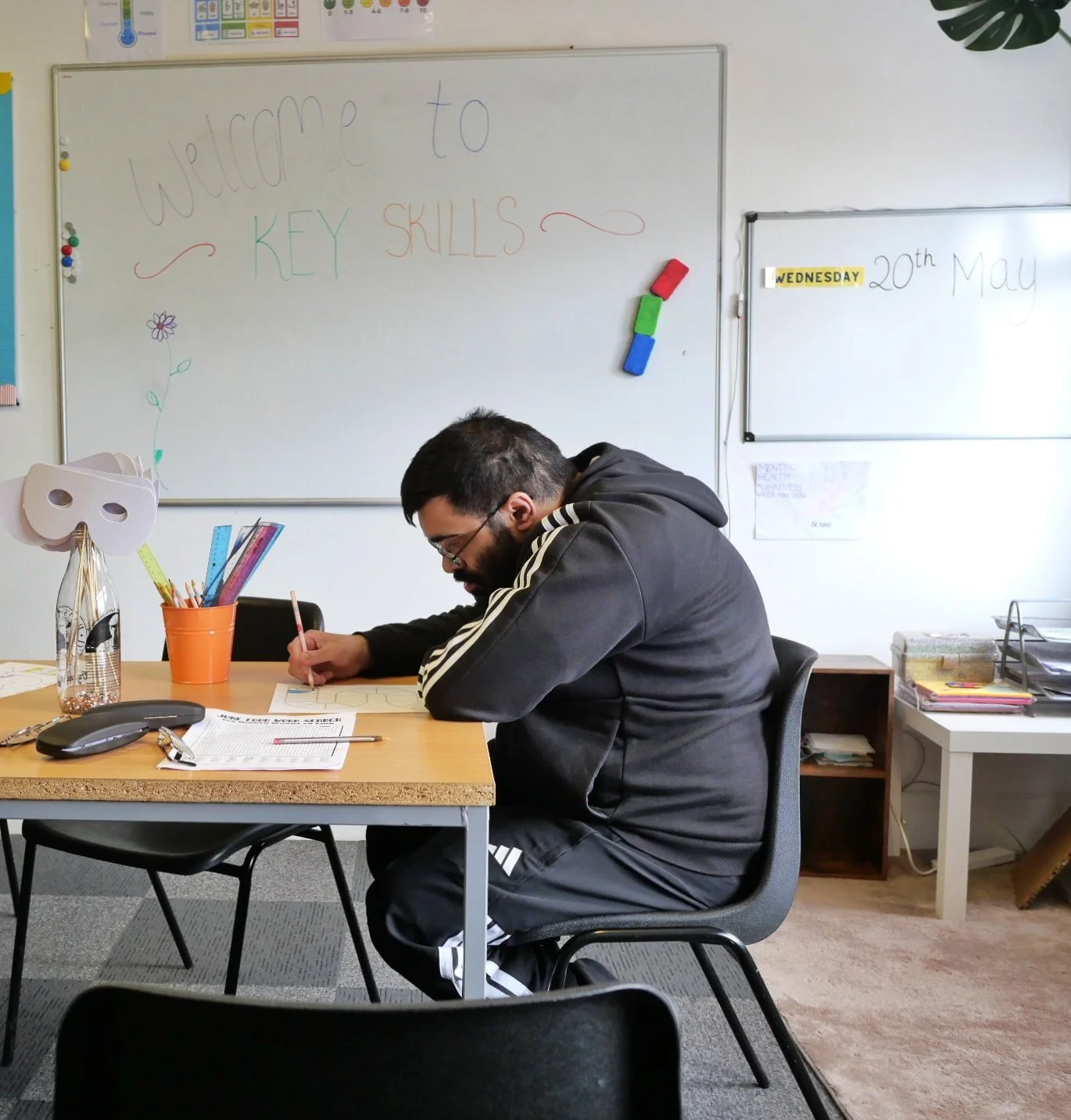 A service user, a man with a learning disability, sits in a classroom at Key. Behind him are whiteboards with colourful writing that says 'Welcome to Key Skills,' and the date 'Wednesday 20th May.'