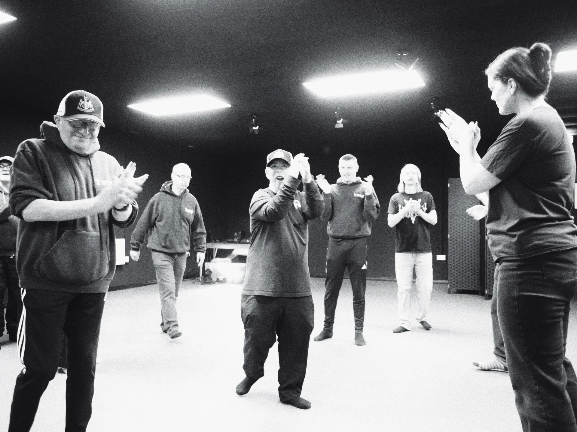 A group of service users in a rehearsal, clapping and dancing in a black box studio at Key, with a woman leading on the right.