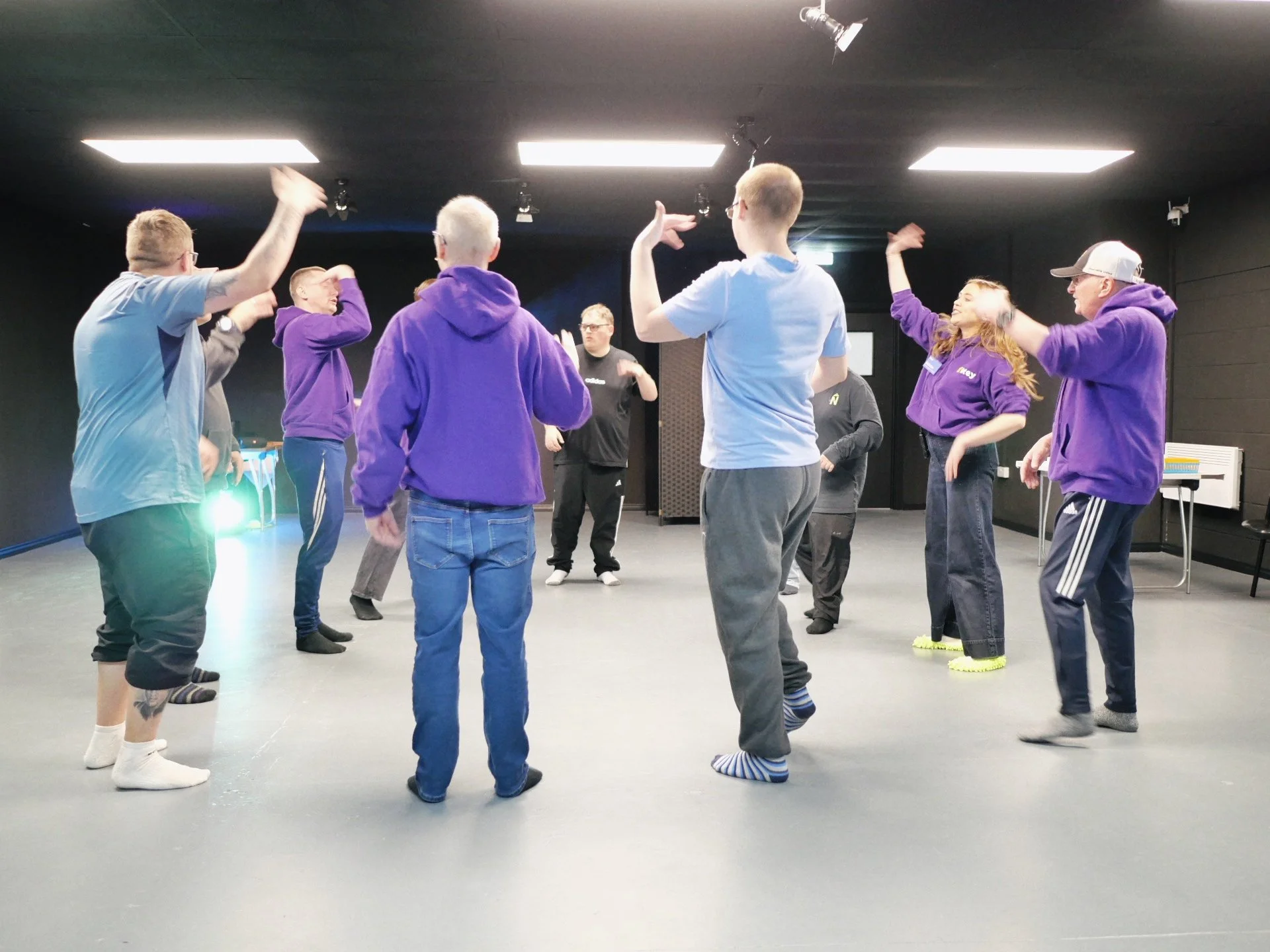 A group of service users with learning disabilities and acquired brain injuries participating in dance and drama in a black box studio at Key, most wearing casual and comfortable clothing with some in purple Key merch hoodies.