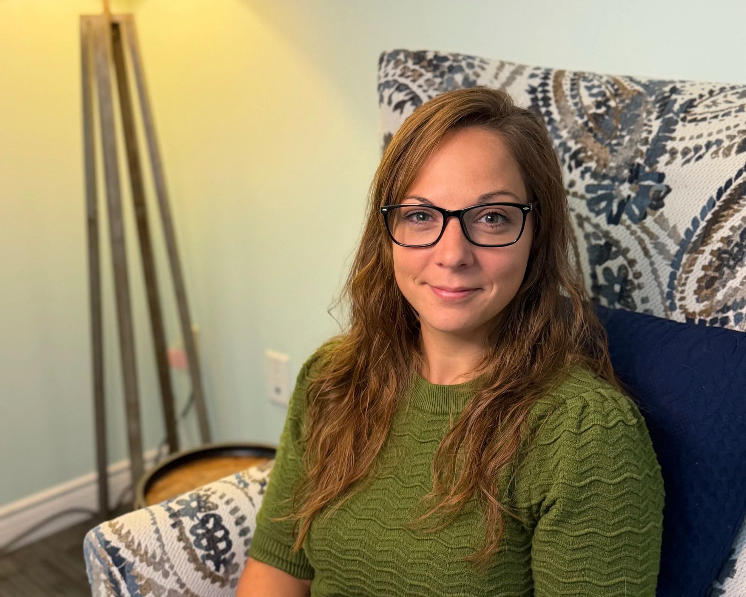 A woman with brown hair, glasses, and a green sweater sitting on a patterned armchair in a living room, smiling at the camera.