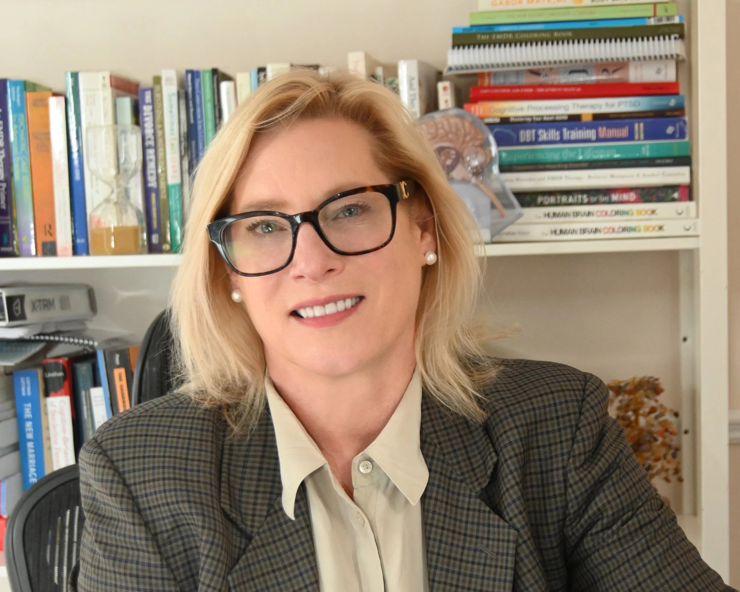 A woman with blonde hair wearing glasses, a checkered blazer, and pearl earrings sitting at a desk with a laptop in front of her. There are books on a bookshelf behind her and a colorful brain model on top of the shelf.