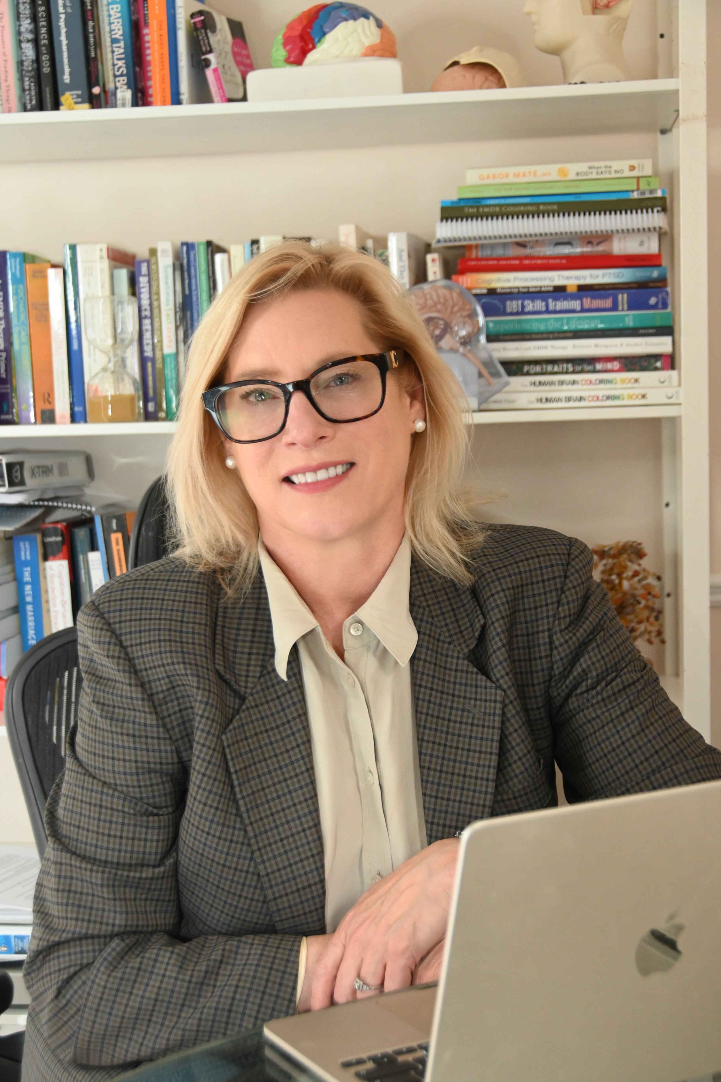 A blonde woman wearing glasses and a plaid blazer, sitting at a desk with a laptop, in an office with bookshelves in the background.