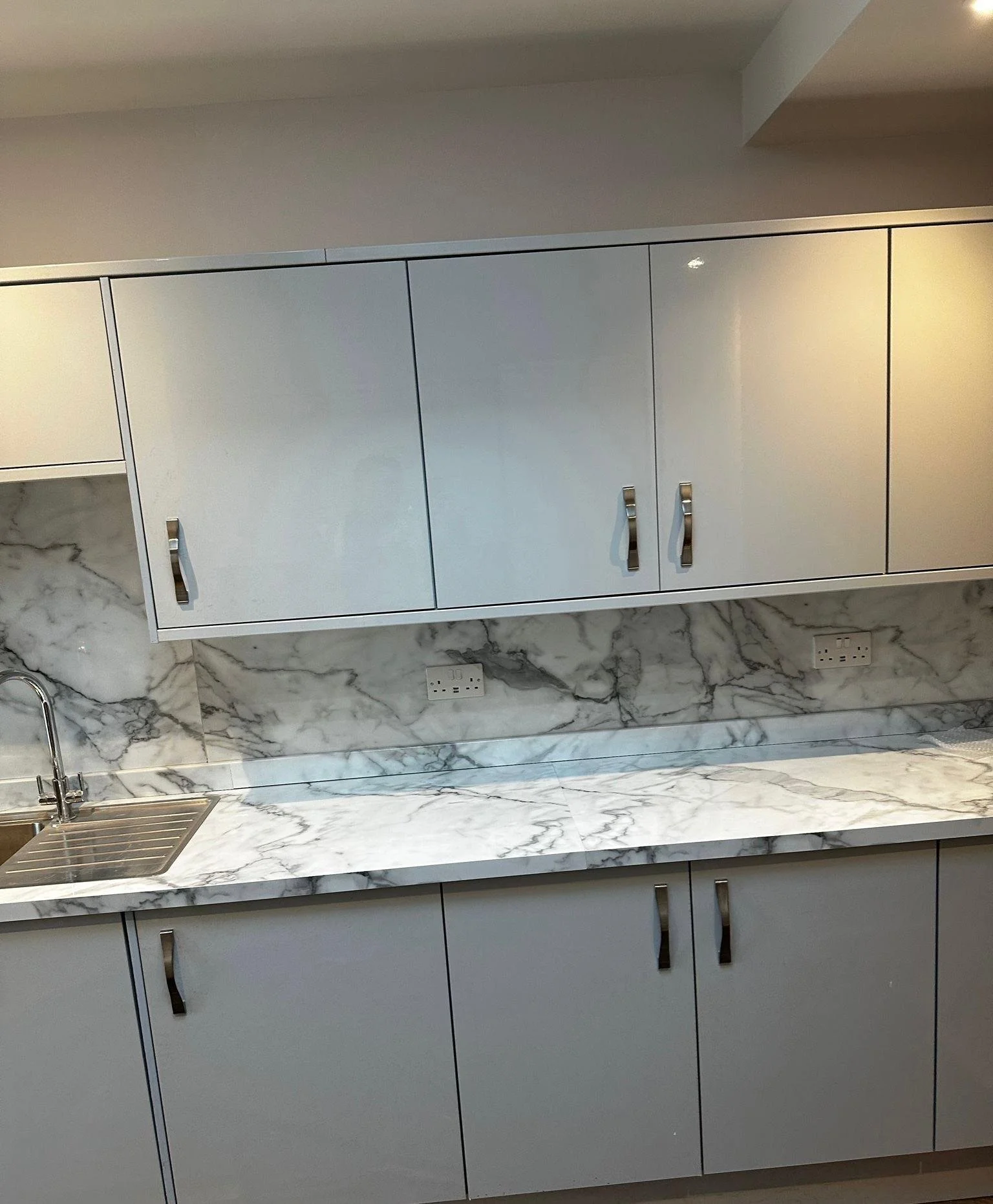 A modern kitchen with grey cabinets, a marble countertop, a stainless steel sink, and electrical outlets on the wall.