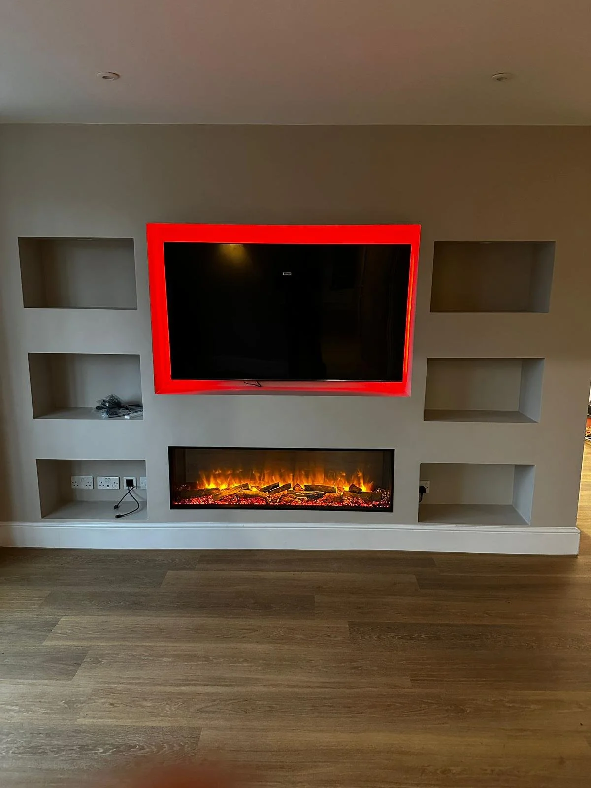 Living room entertainment wall with a flat-screen TV framed by red lights, an electric fireplace below, and empty wall niches.