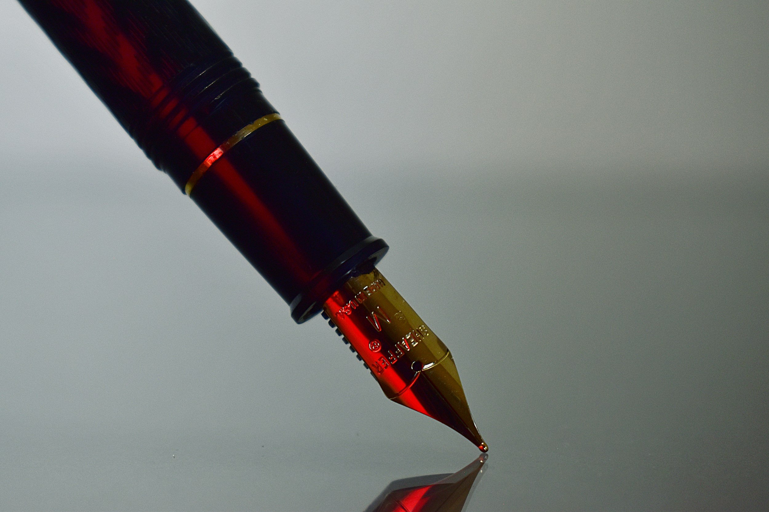 Close-up of a red and black fountain pen with gold accents, positioned at an angle against a gray background, with its reflection on a glossy surface.