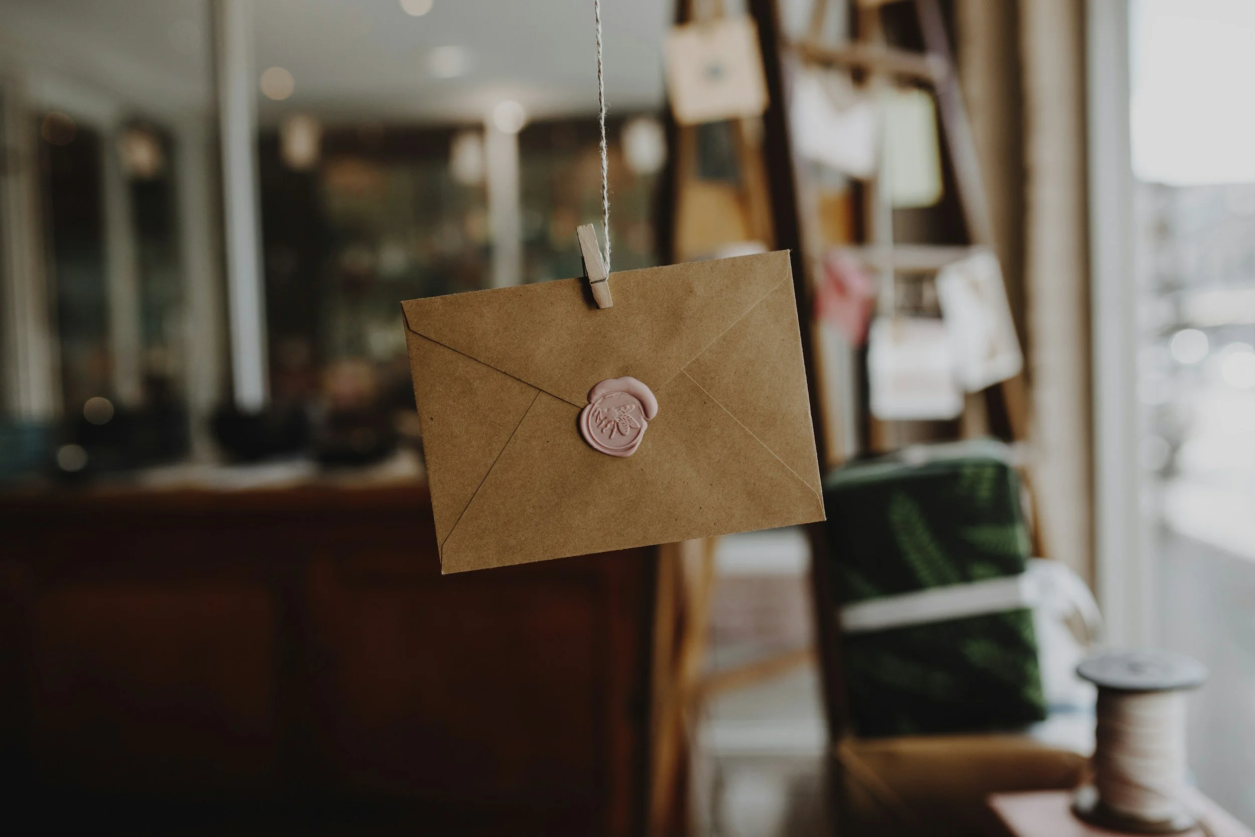 Brown envelope with pink wax seal hanging from a string with a clothespin in a cozy indoor shop or café setting.