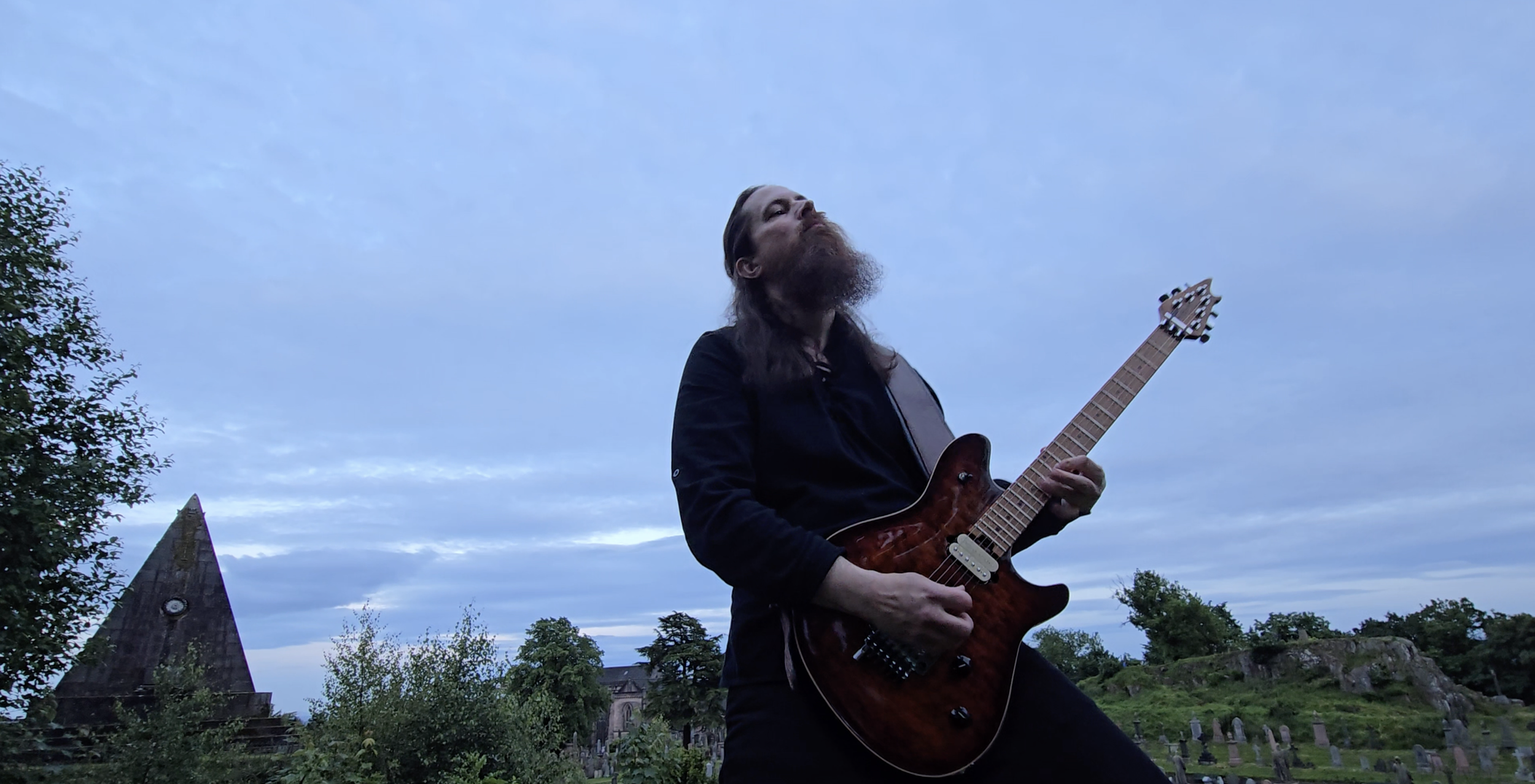 A man with long hair, beard, and black clothing playing an electric guitar outdoors during dusk, with a cloudy sky and trees in the background.