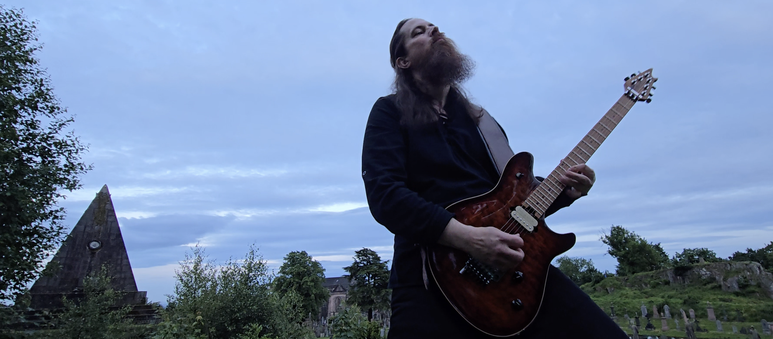 A man with long hair and a beard playing an electric guitar outdoors in a grassy area with trees and gravestones, under a cloudy sky.