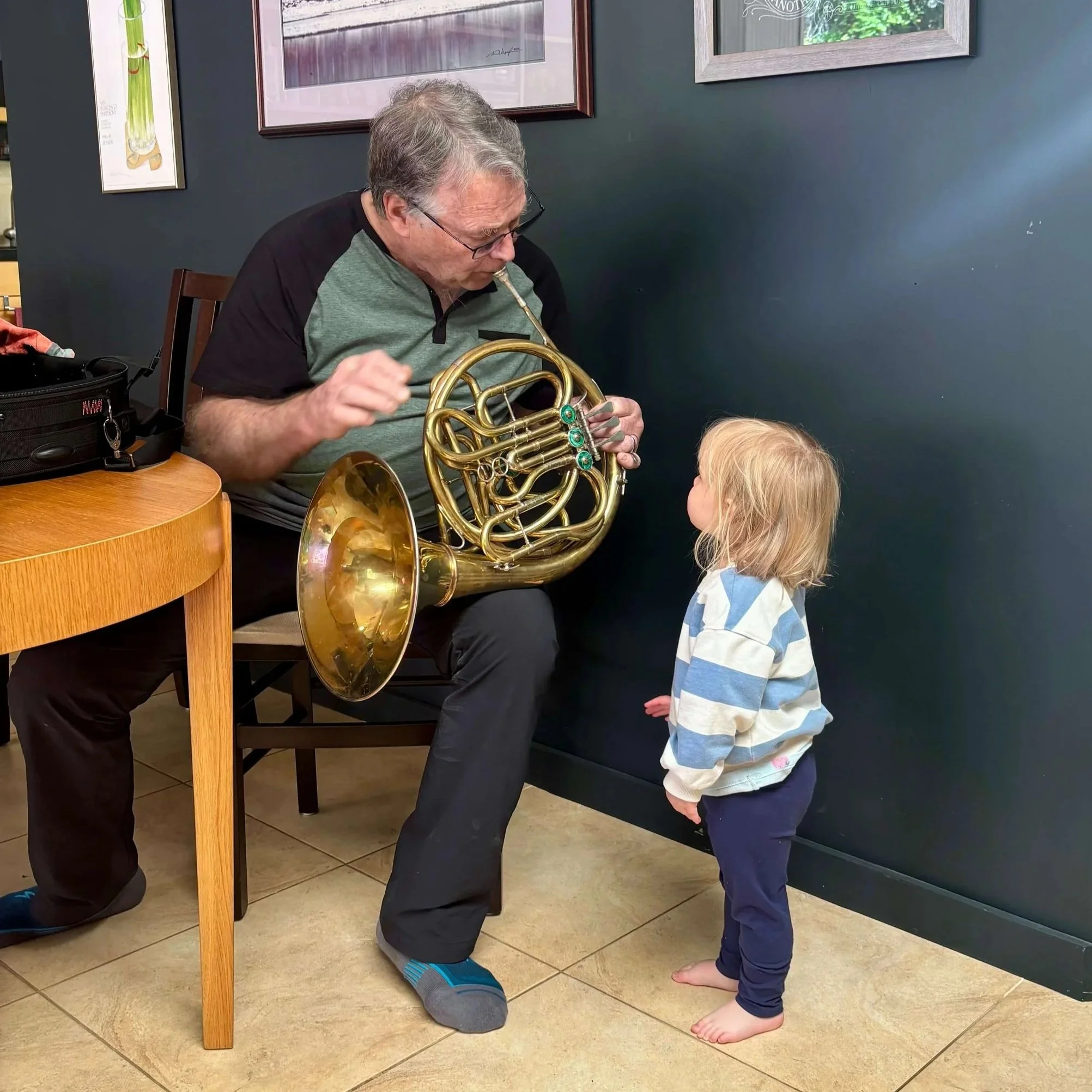 Bruce Roberts, hornmaker, sitting on a chair, holding a French horn, and engaging with a young child who is standing barefooted on tiled floor. They are facing each other, and the man appears to be explaining something about the French honr