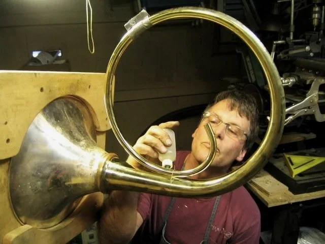 Bruce Roberts working on a French horn, in a workshop with tools and equipment in the background.