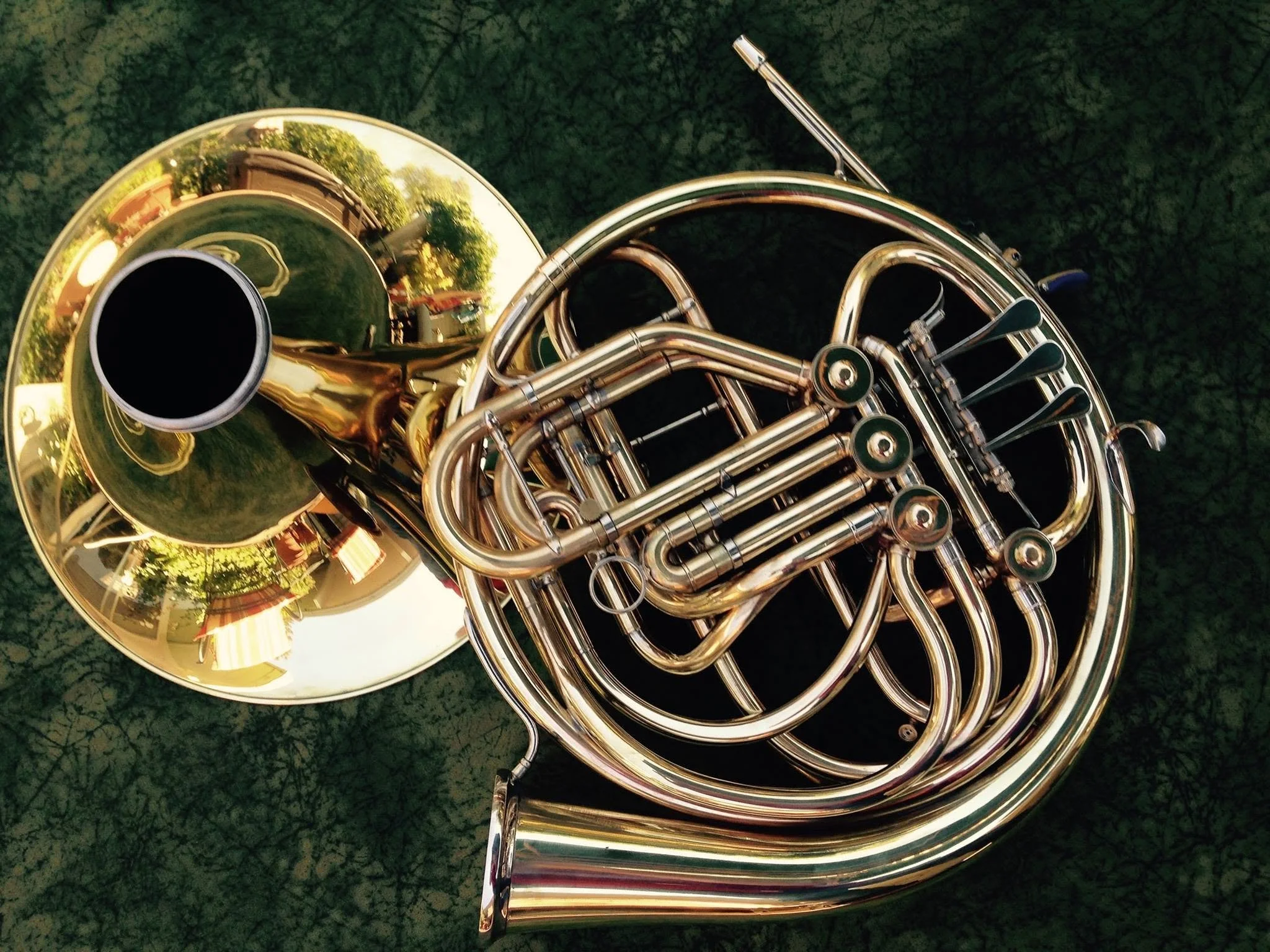 A shiny brass French horn resting on a dark green patterned surface, with its bell and intricate tubing visible.