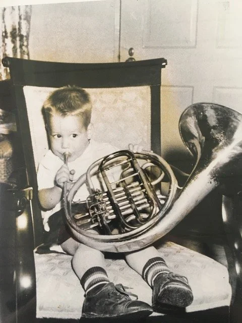 An antique black and white photo of a young boy sitting on a chair with a little French horn resting on his lap, wearing striped socks and shoes, and holding the mouthpiece to his lips.
