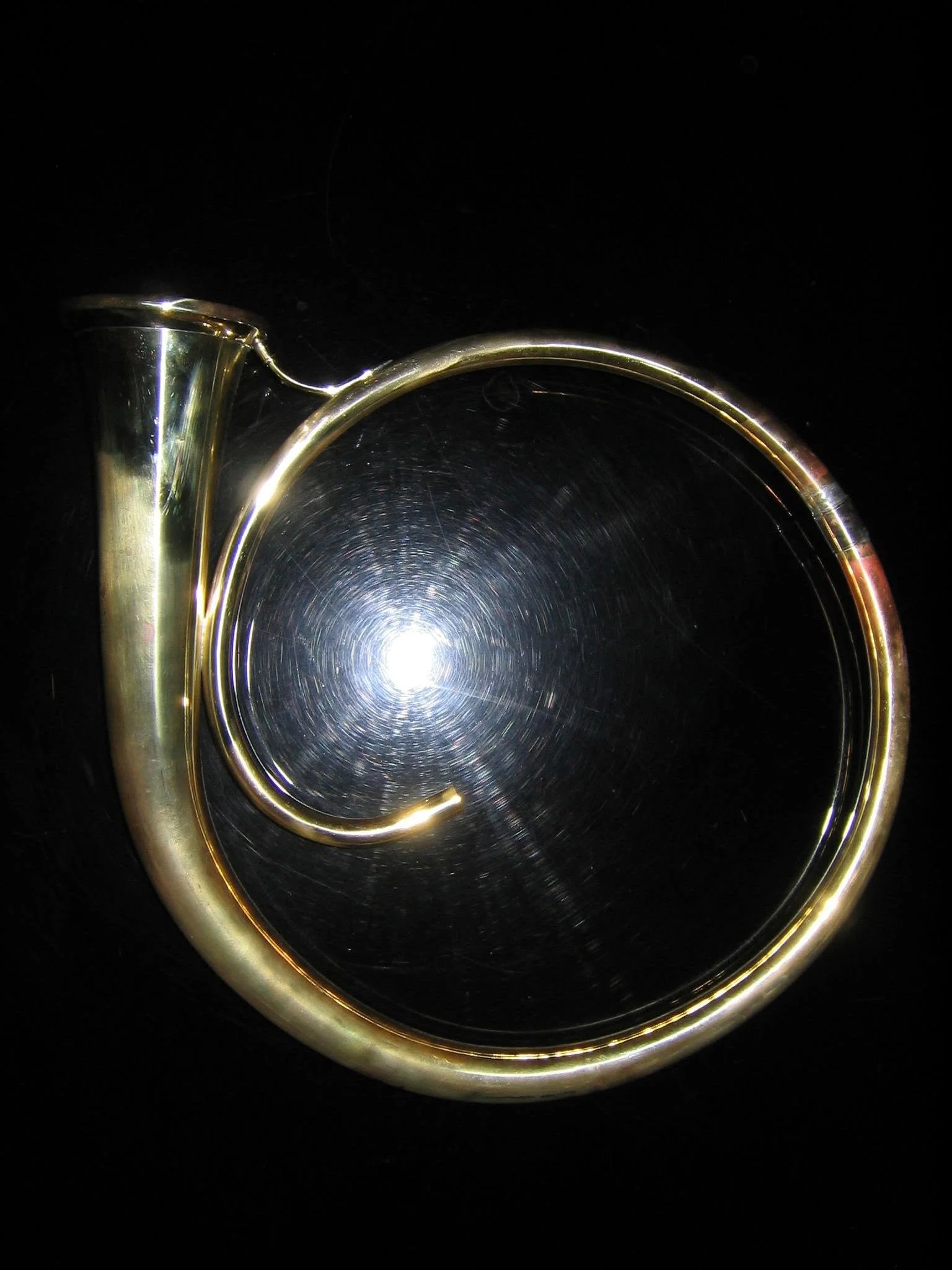 Close-up of a shiny brass French horn on a reflective surface against a black background, resembling part of a musical instrument or decorative item.
