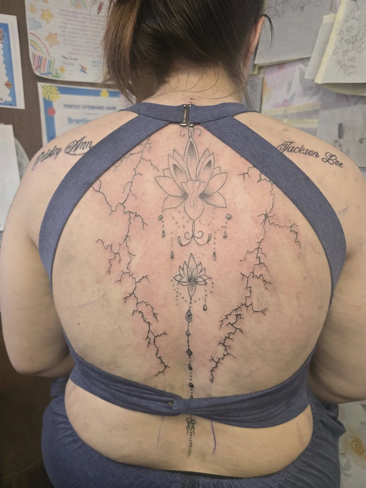 A person with a large back tattoo featuring a line of lotus flowers connected by a decorative chain, with the person's back facing the camera and wearing a dark gray top with straps.