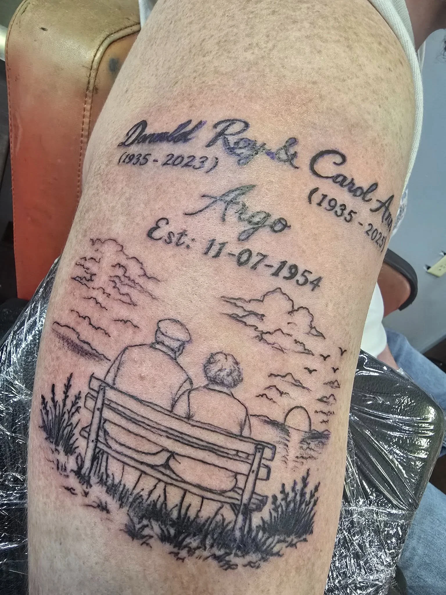 A black ink tattoo on a person's upper arm showing a couple sitting on a park bench, with a background of clouds and birds in the sky. The tattoo includes text and dates.