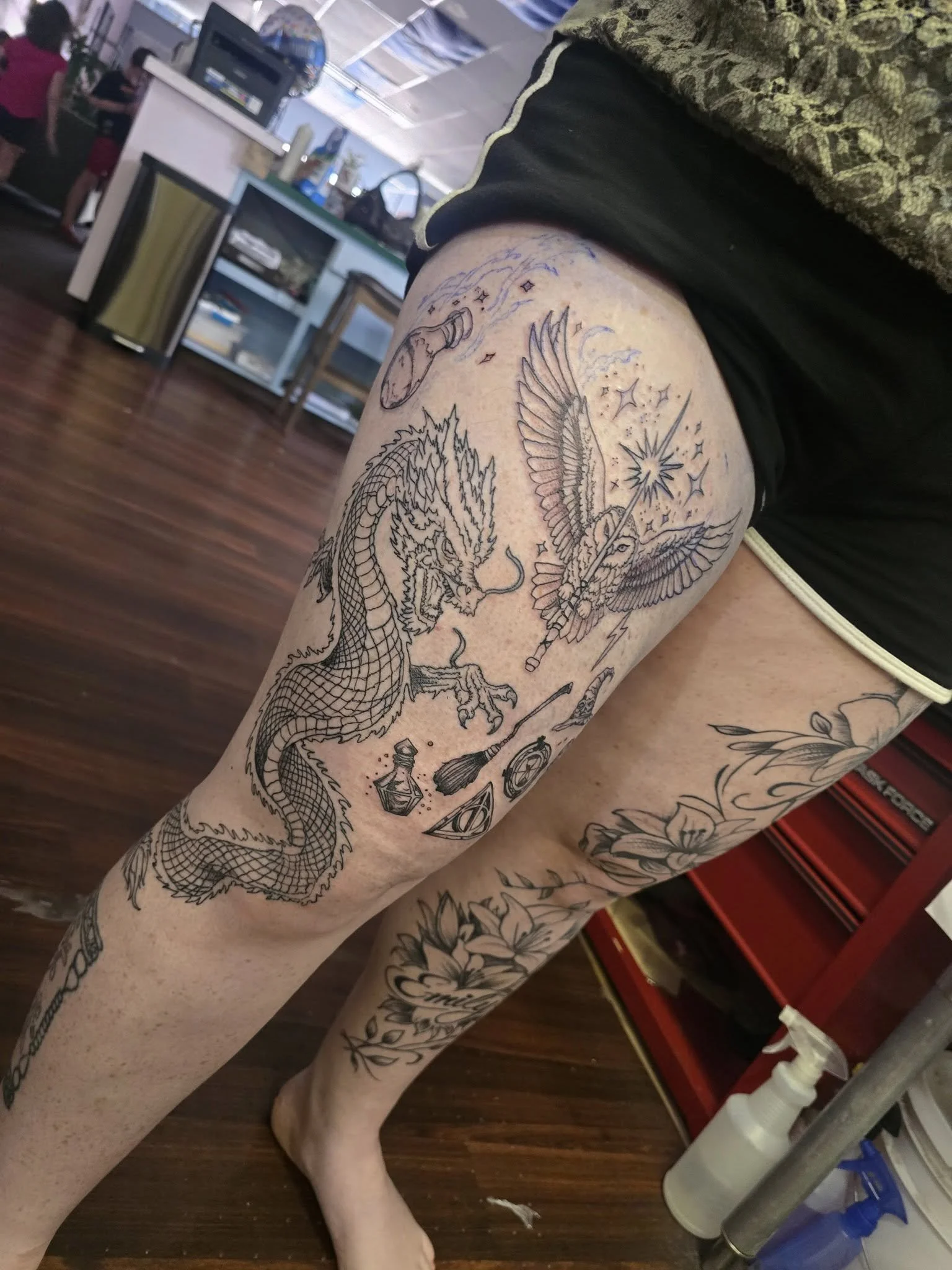 A person's upper leg with multiple tattoos, including a dragon, a winged creature holding a wand, and various mystical symbols, inside a tattoo studio.