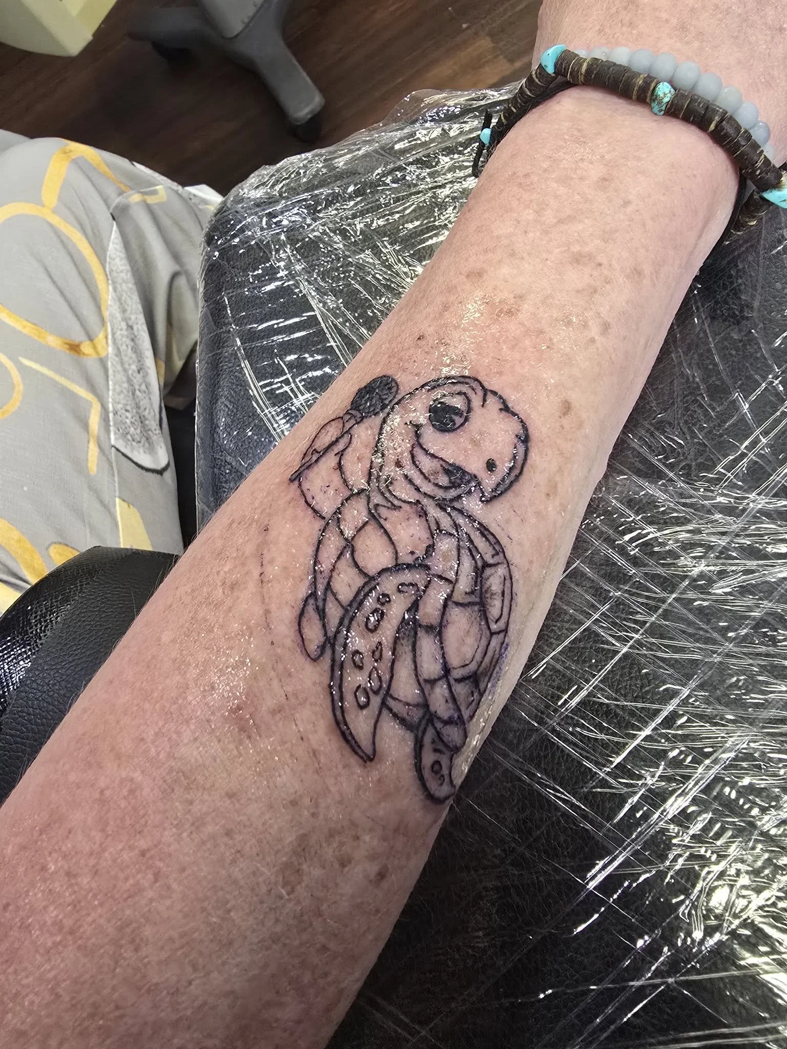 A person's forearm with a tattoo outline of a cartoon dog, possibly Snoopy, on the skin. The arm has a beaded bracelet with turquoise and gray beads. The surrounding area is covered with plastic wrap, likely due to fresh tattoo work.