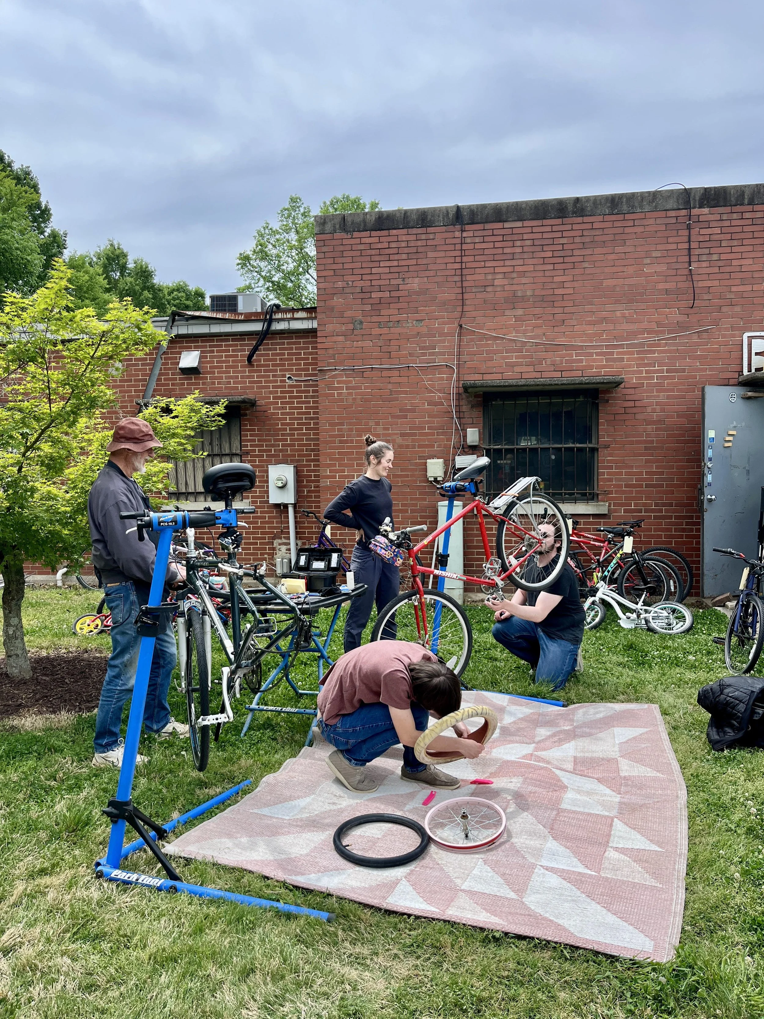 Group of people working on fixing and assembling bicycles outside in a grassy area with a brick building in the background. Some are kneeling and others are standing, with tools, bike parts, and a large carpet on the ground.