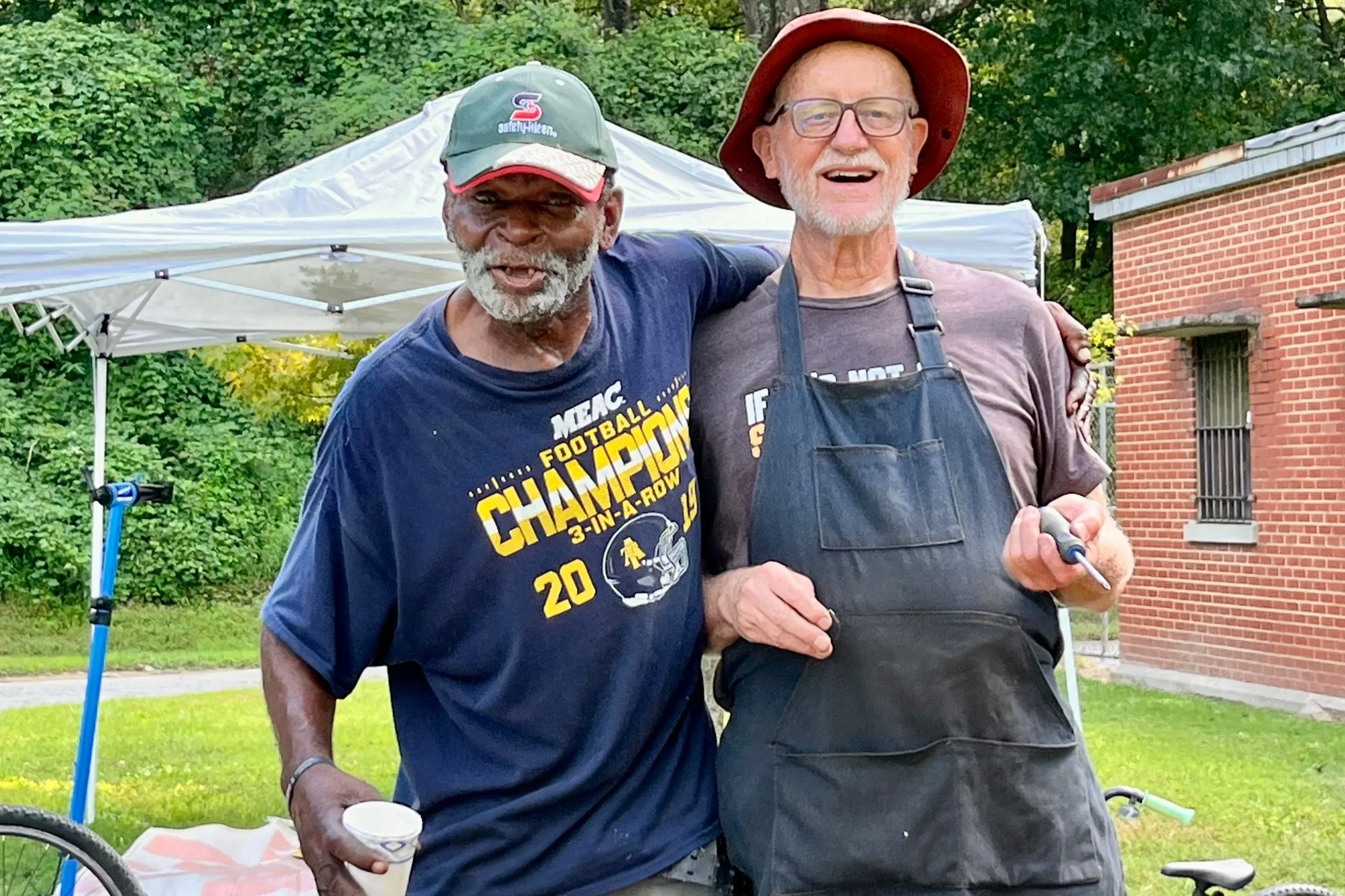 Two elderly men smiling and posing outdoors, one wearing a baseball cap and a blue T-shirt with 'Championship' written on it, and the other in a wide-brimmed hat and apron, standing in front of a white canopy tent, with green trees and a brick building in the background.