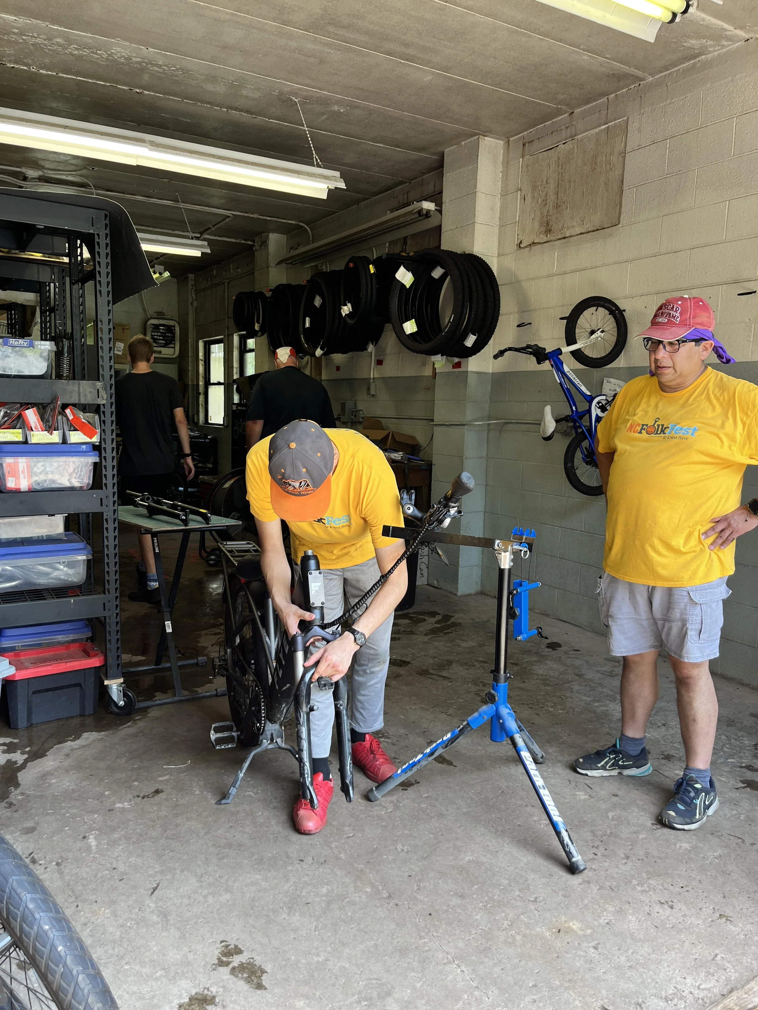 Three people working in a bike shop; one person is assembling a bicycle, another is standing to the right wearing a yellow MC Folk Fest shirt and a red hat, and a third person is in the background near the wall. Bike tires are hung on the wall, and shelves with storage bins are visible.