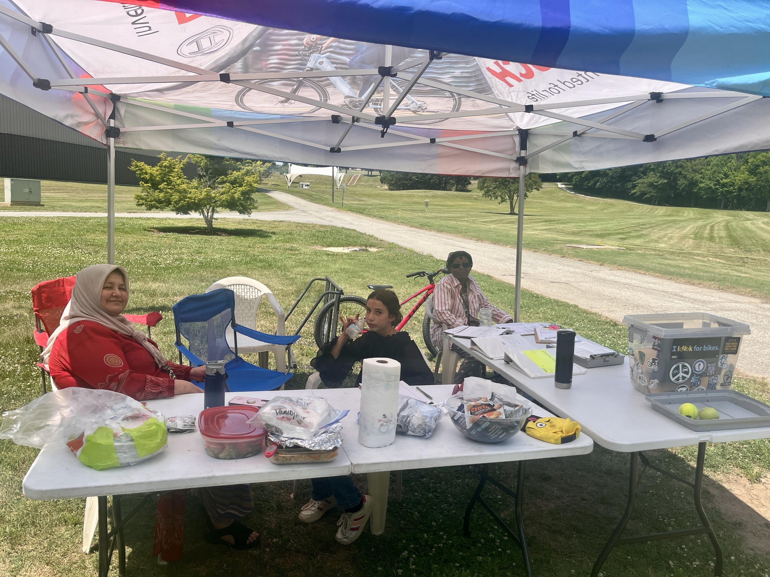 A group of people sitting around a table under a canopy outdoors on a grassy area. The table has various food items, drinks, papers, and other supplies. Two women and a child are visible, with bicycles and a pathway leading into a neighborhood in the background.