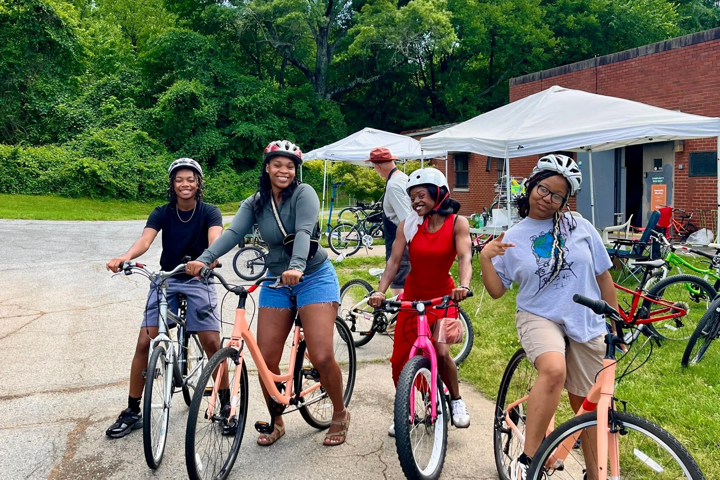 Group of five women on bicycles smiling outdoors near tents and a brick building with trees in the background.