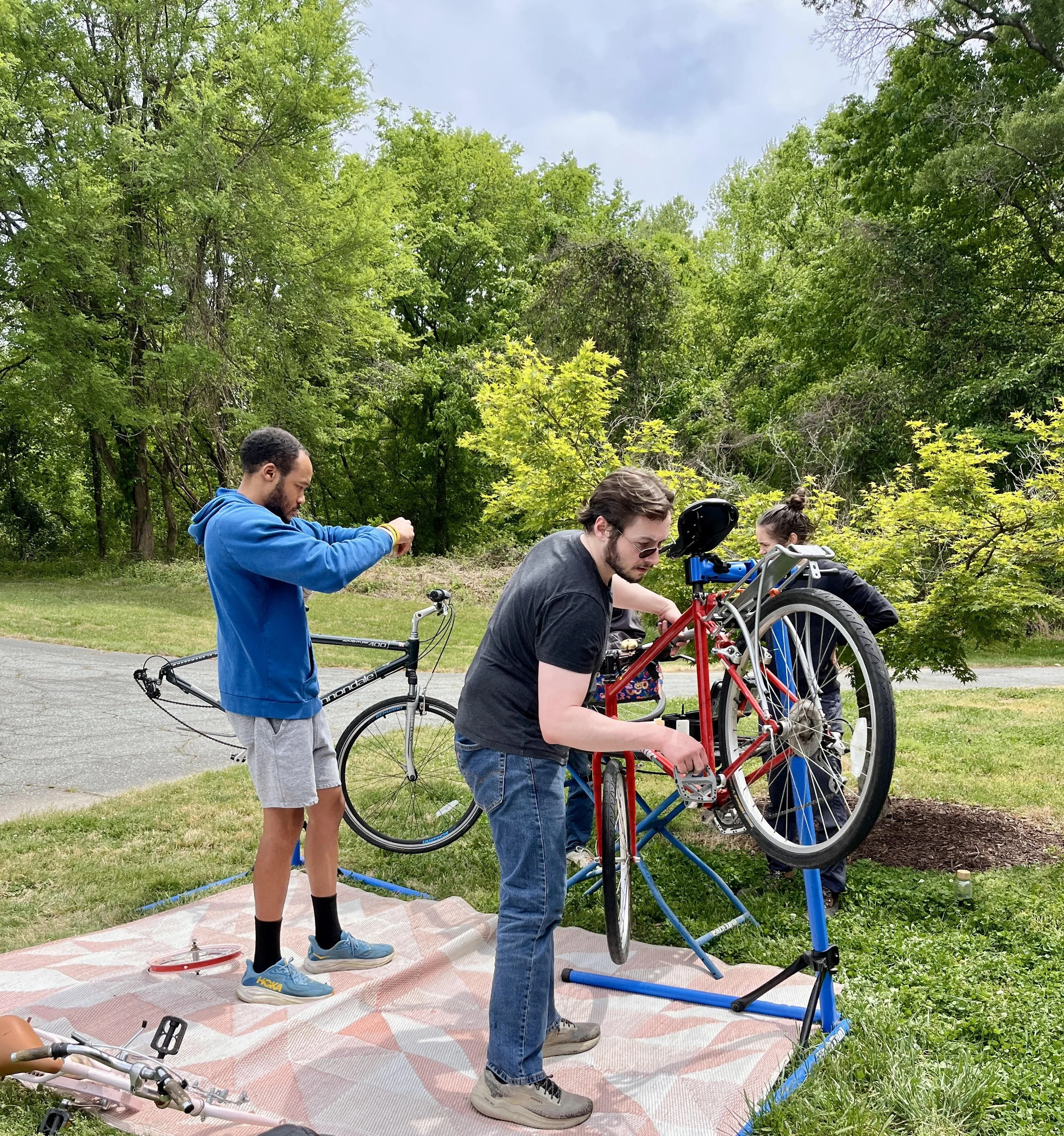 Group of people repairing and assembling a bicycle outdoors on a grassy area with trees in the background. One person is working on the bicycle while others are observing or taking photos.